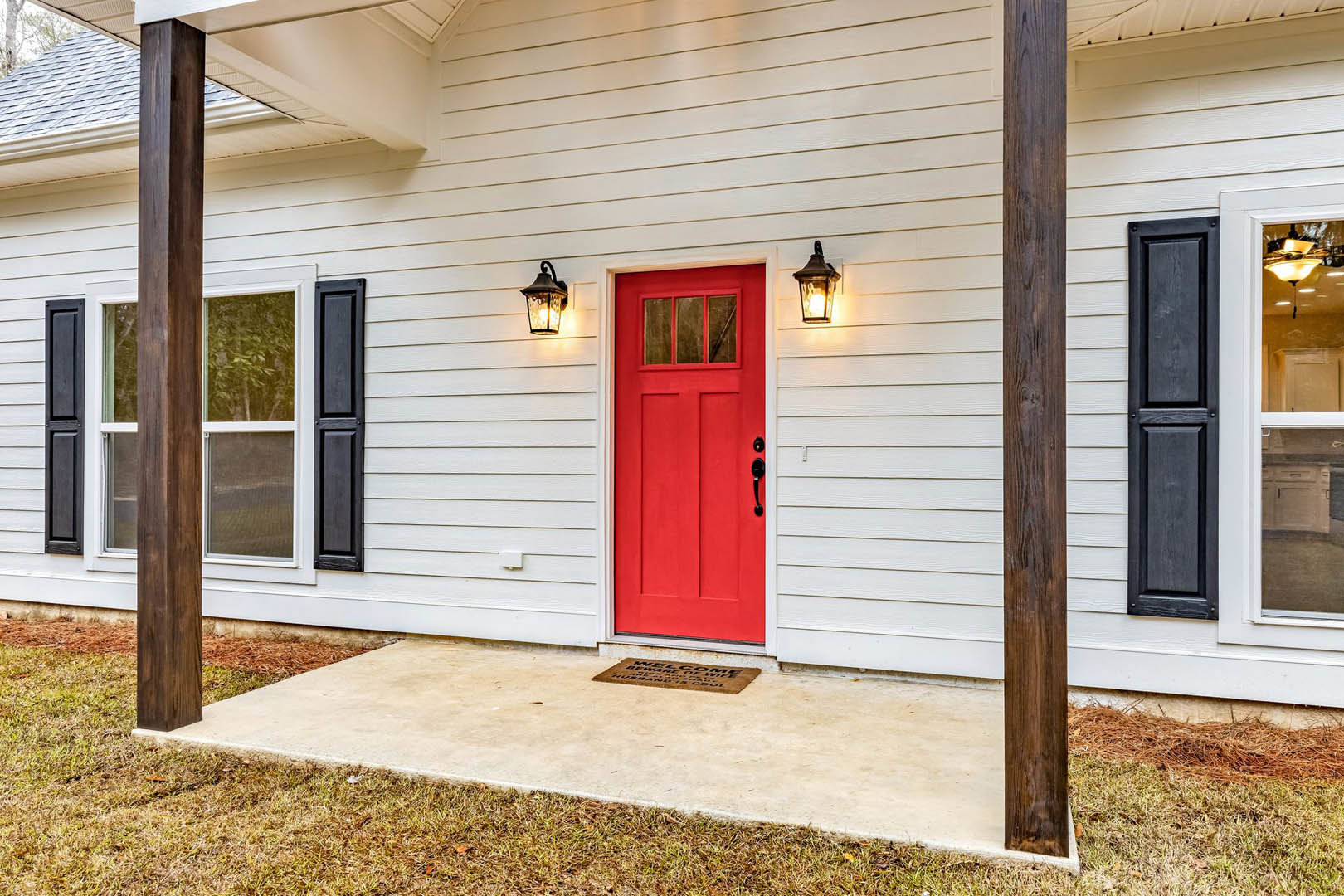 Red front door with black hardware on white siding, brown welcome mat on concrete porch, adjacent window with white trim, green grass bordering entry walkway.