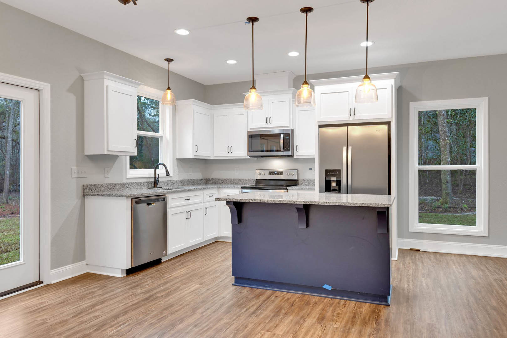 Modern kitchen featuring a central island with quartz countertop, stainless steel appliances including a built-in microwave, white cabinetry, large window framing a tree trunk, and