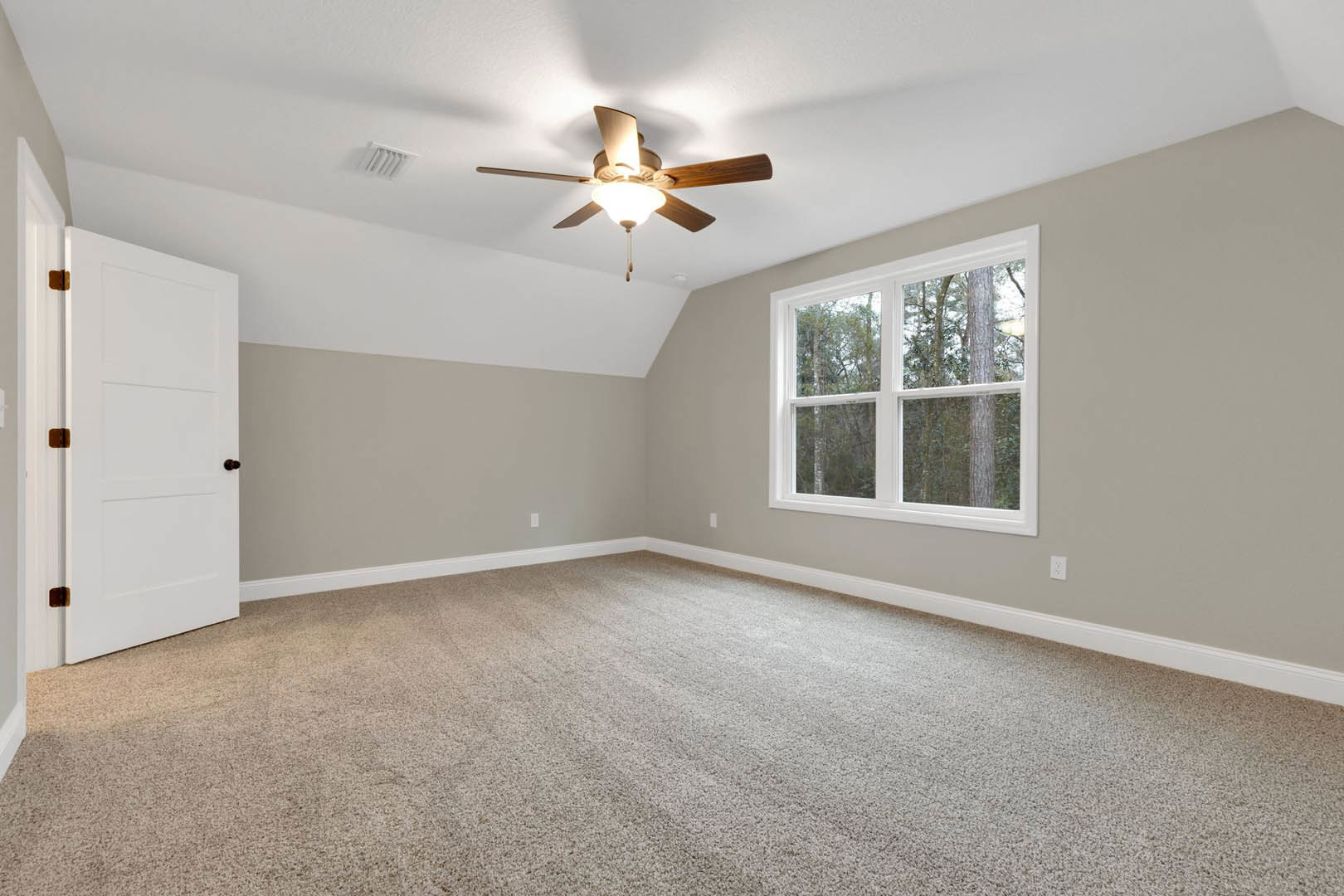 Carpeted room with white walls, ceiling fan with light fixture, multiple windows showing trees outside, and a white door