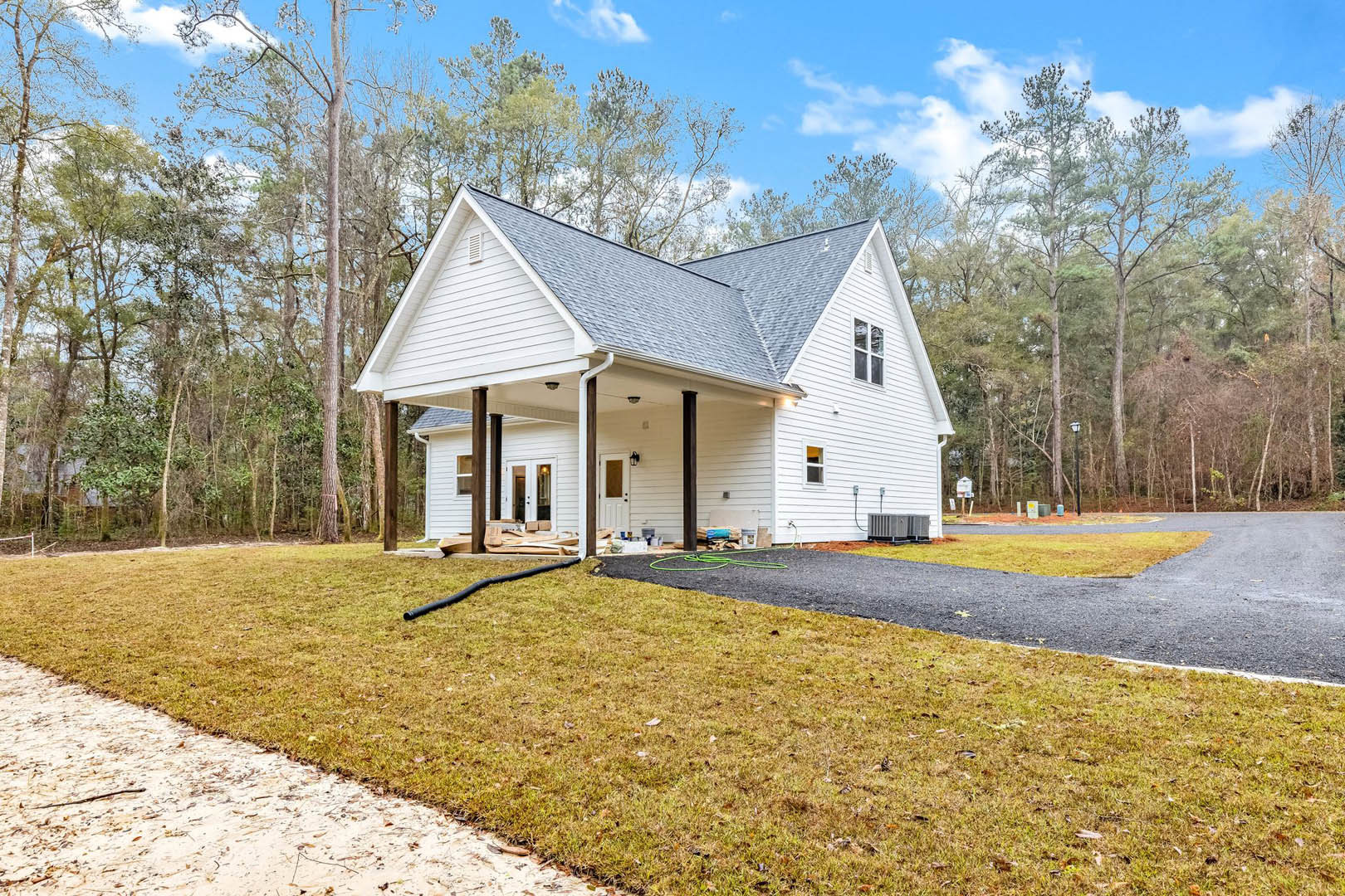 White siding house with covered porch, concrete driveway, green lawn, black garden hose, and mature trees in the background
