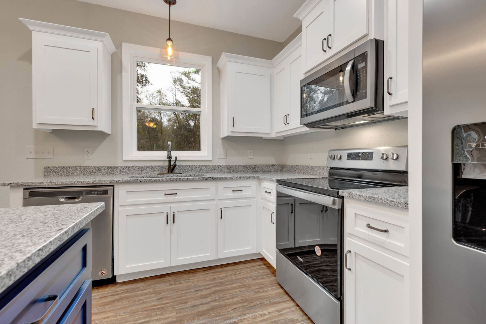 White kitchen with granite countertops, stainless microwave, pendant ceiling light, window overlooking trees, wood flooring, and white cabinetry with metal handles.