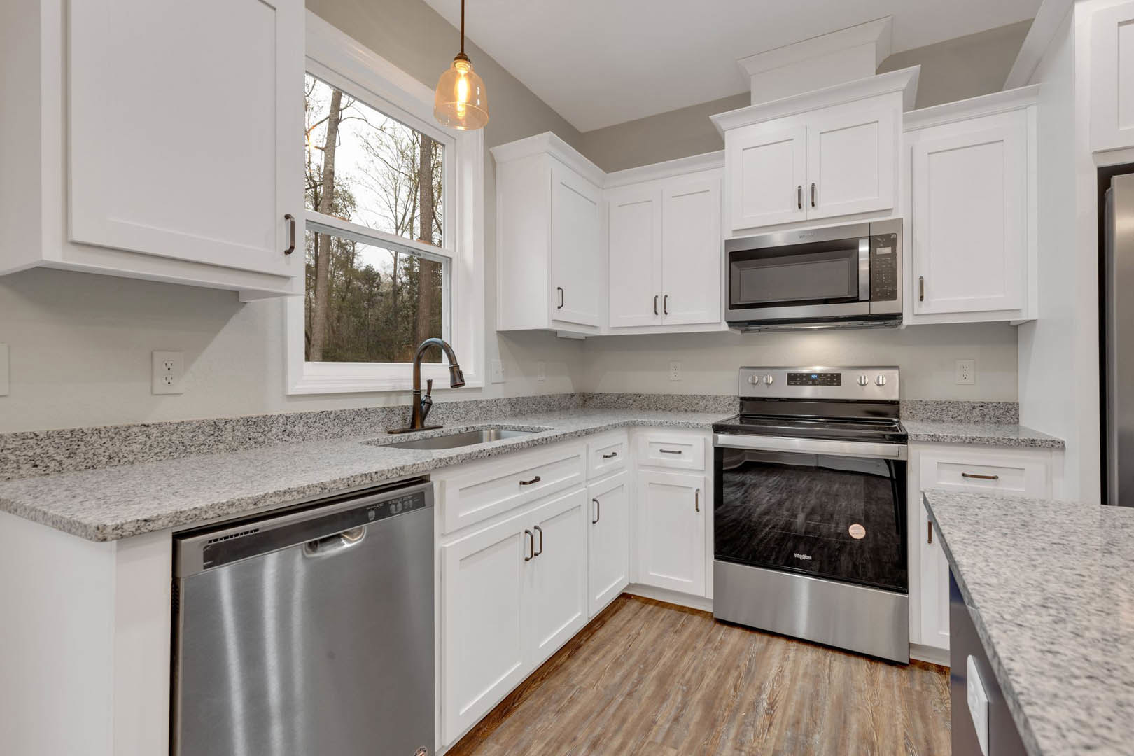 White kitchen cabinets, granite countertops, stainless steel dishwasher, oven, and microwave, stove with controls visible, window overlooking green trees