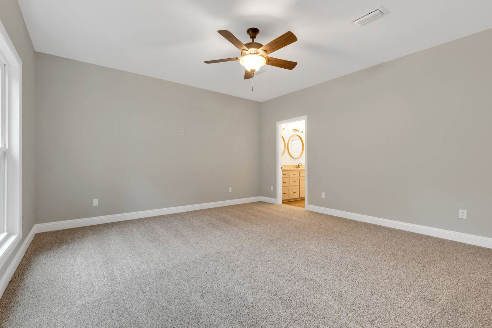 Carpeted bedroom with white ceiling fan, open door leading to bathroom with vanity and mirror, neutral walls, white trim, and visible cabinet drawer.