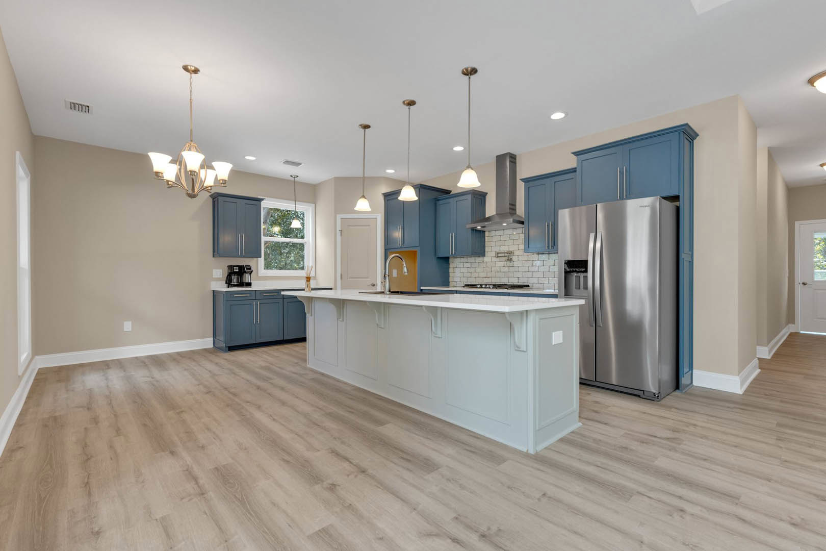 Blue shaker cabinets, stainless steel refrigerator, white kitchen island, wood flooring, white walls, modern chandelier, window with white frame and light fixture