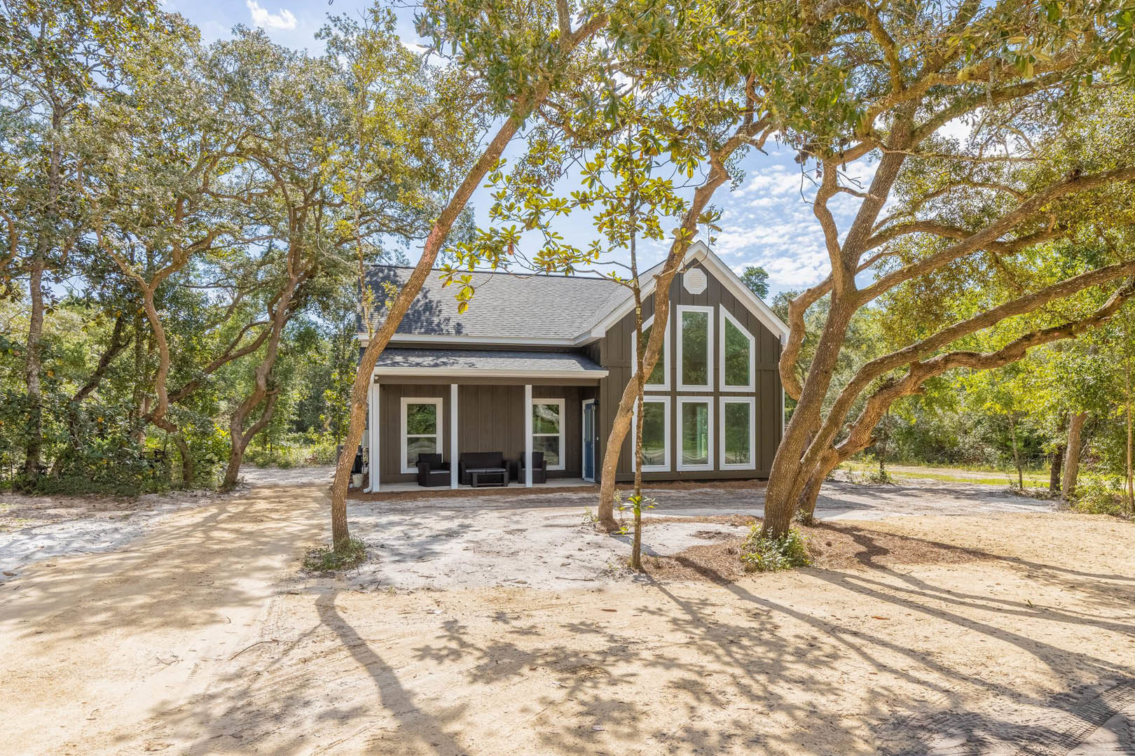 Two-story home surrounded by mature trees, white-framed windows, yellow leaves scattered on roof, outdoor seating area with couch and table on dirt ground