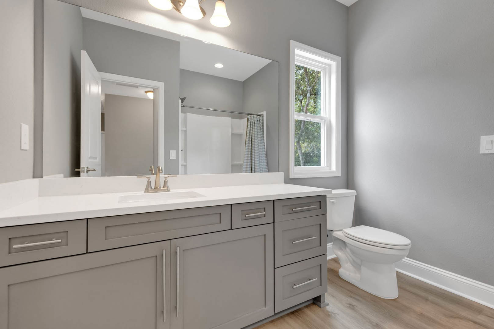 Bathroom with white toilet, rectangular sink on stone countertop, striped curtain beside window showing trees, wall-mounted light fixture, neutral tile walls and cabinetry