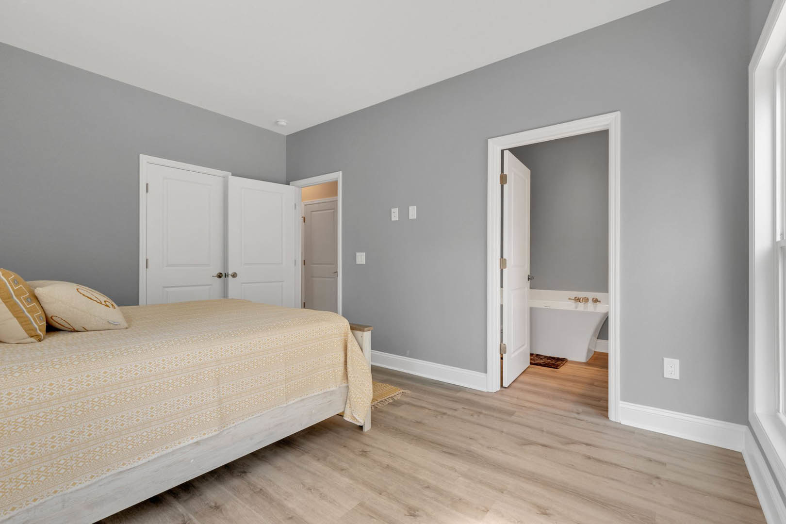 Bedroom with light wood flooring, upholstered bed with white bedding and pillows, freestanding bathtub near a white door with metal handle, neutral walls and minimalist decor.