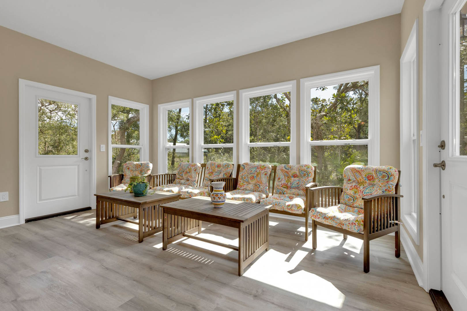 Living room with wood flooring, a wooden table topped with a vase, colorful upholstered chairs with wooden frames, white door with glass window, neutral walls, and natural light