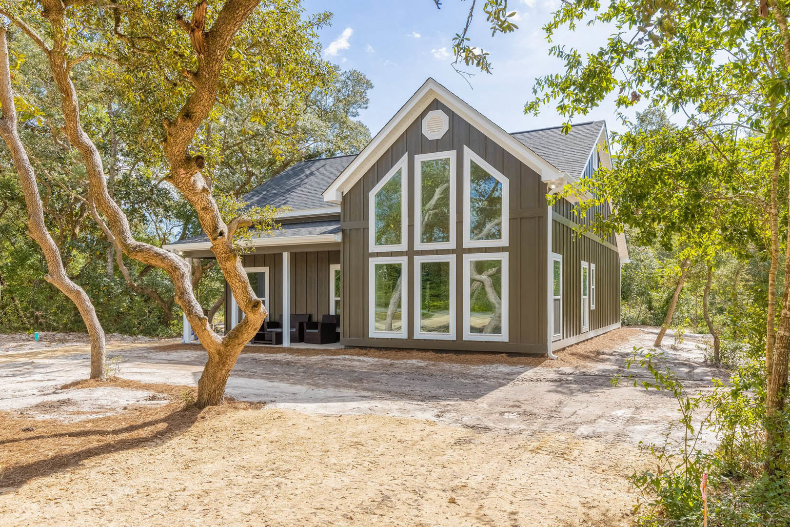 Gray-sided custom home with multiple white-framed windows, surrounded by mature trees and greenery, white vent visible on exterior wall, tree trunk in foreground.
