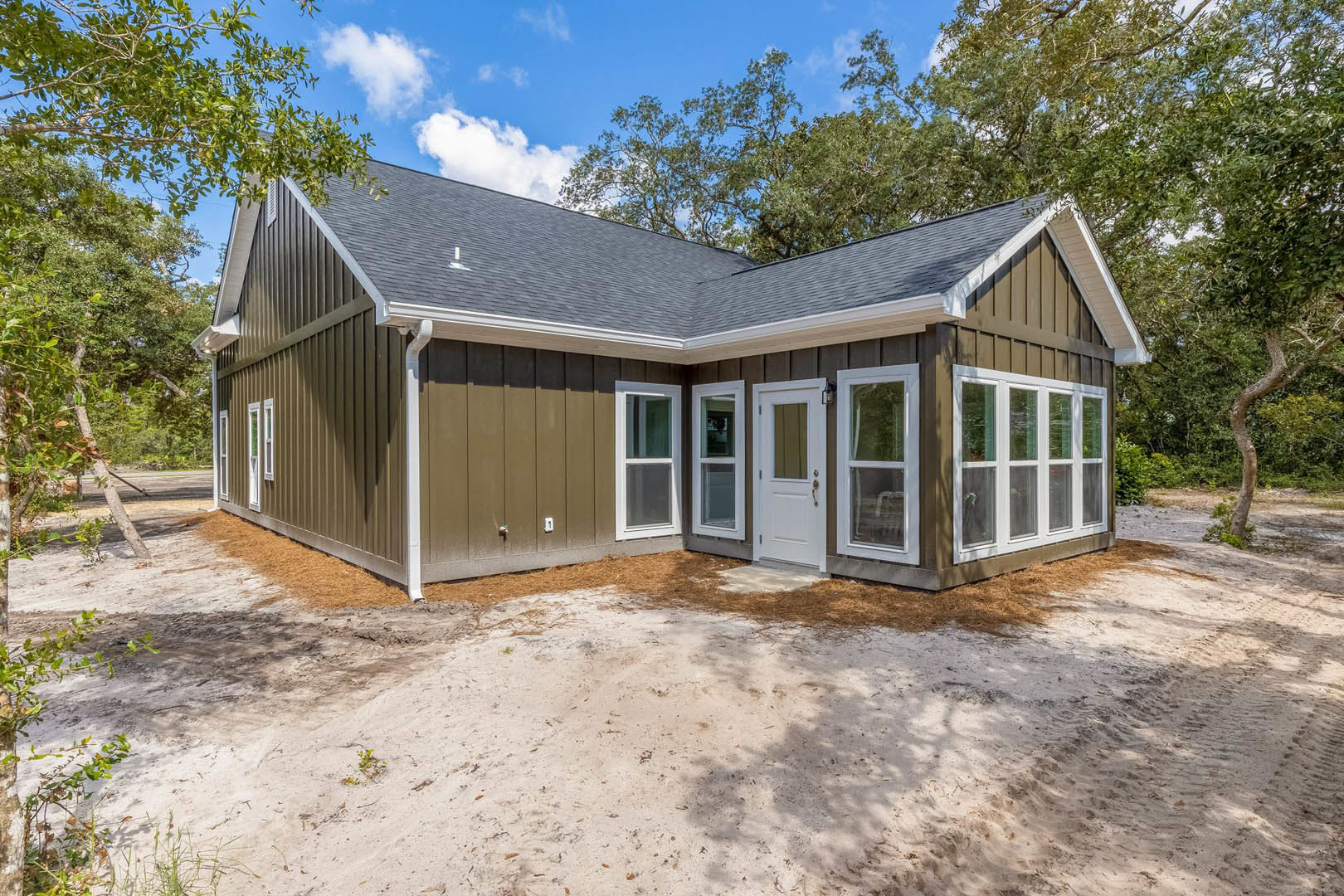 Two-story house surrounded by mature trees, white front door and windows with white frames, dirt ground in foreground, cloudy sky above