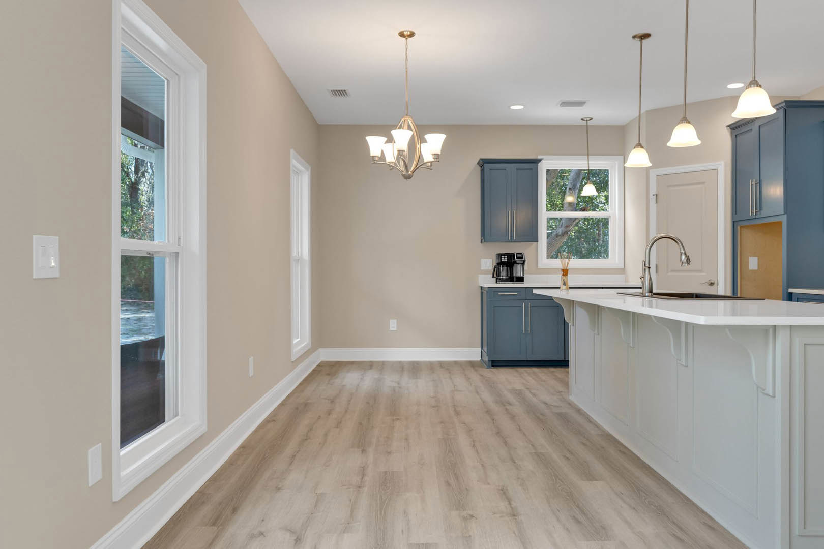 Kitchen with hardwood flooring, blue cabinetry, white light switch on wall, white-framed window showing trees, and tiled backsplash.