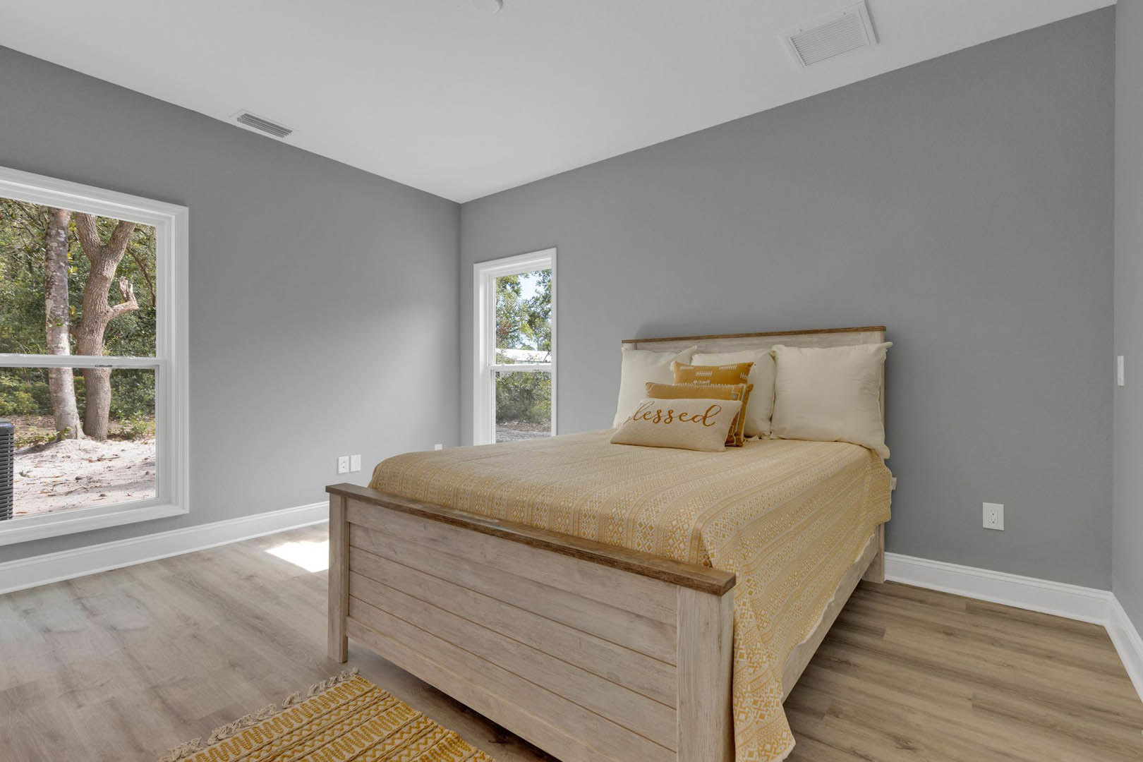 Queen bed with white linens and assorted pillows, patterned area rug on hardwood floor, large window with tree view, neutral walls, minimal decor