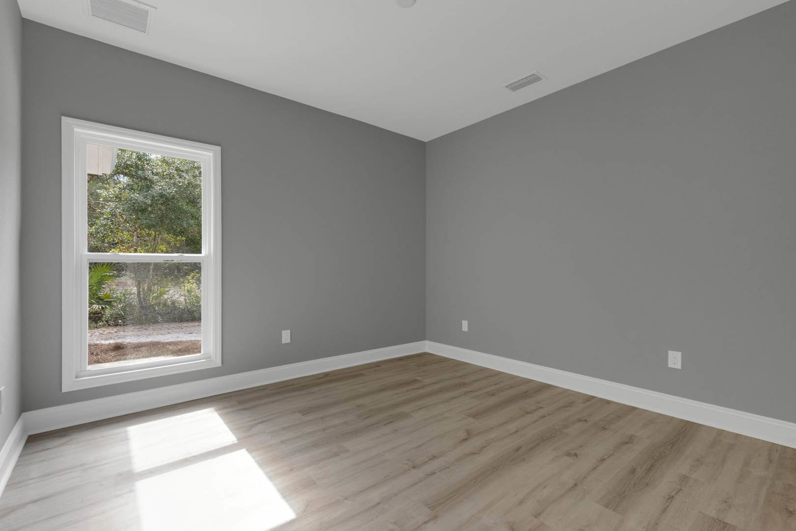 Sunlit room featuring large window overlooking trees, wood flooring with white baseboards, white plaster walls, and a white vent near the floor.