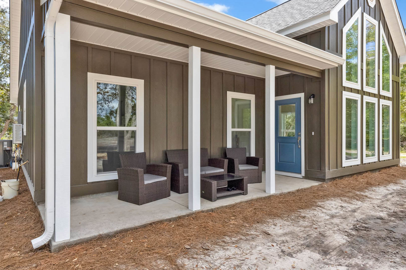 Front porch with wicker chairs and cushions, blue door, white-framed window, small table, and white bucket with black handle on light-colored siding