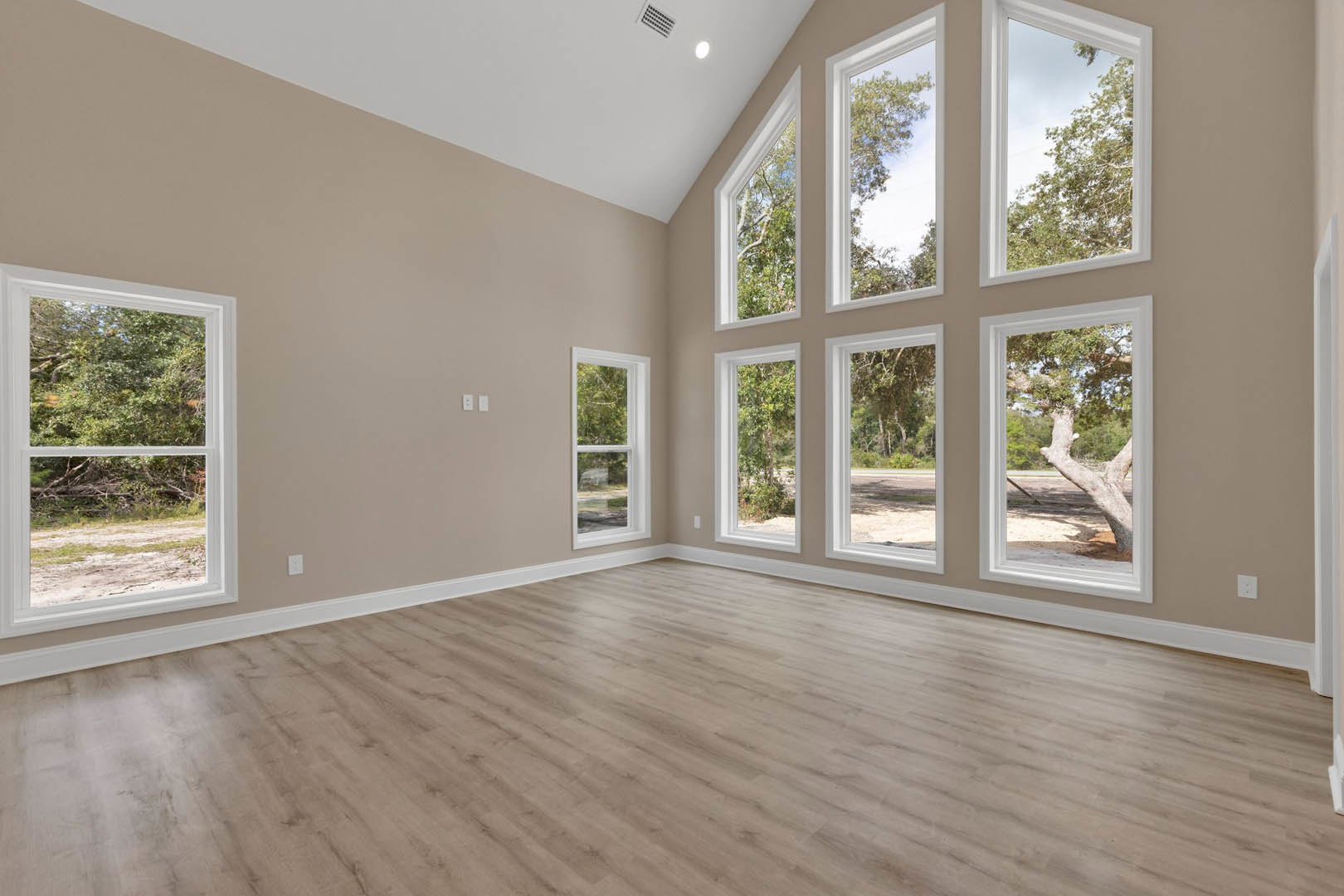 Spacious room featuring hardwood floors, multiple large white-framed windows, and views of leafy trees outside