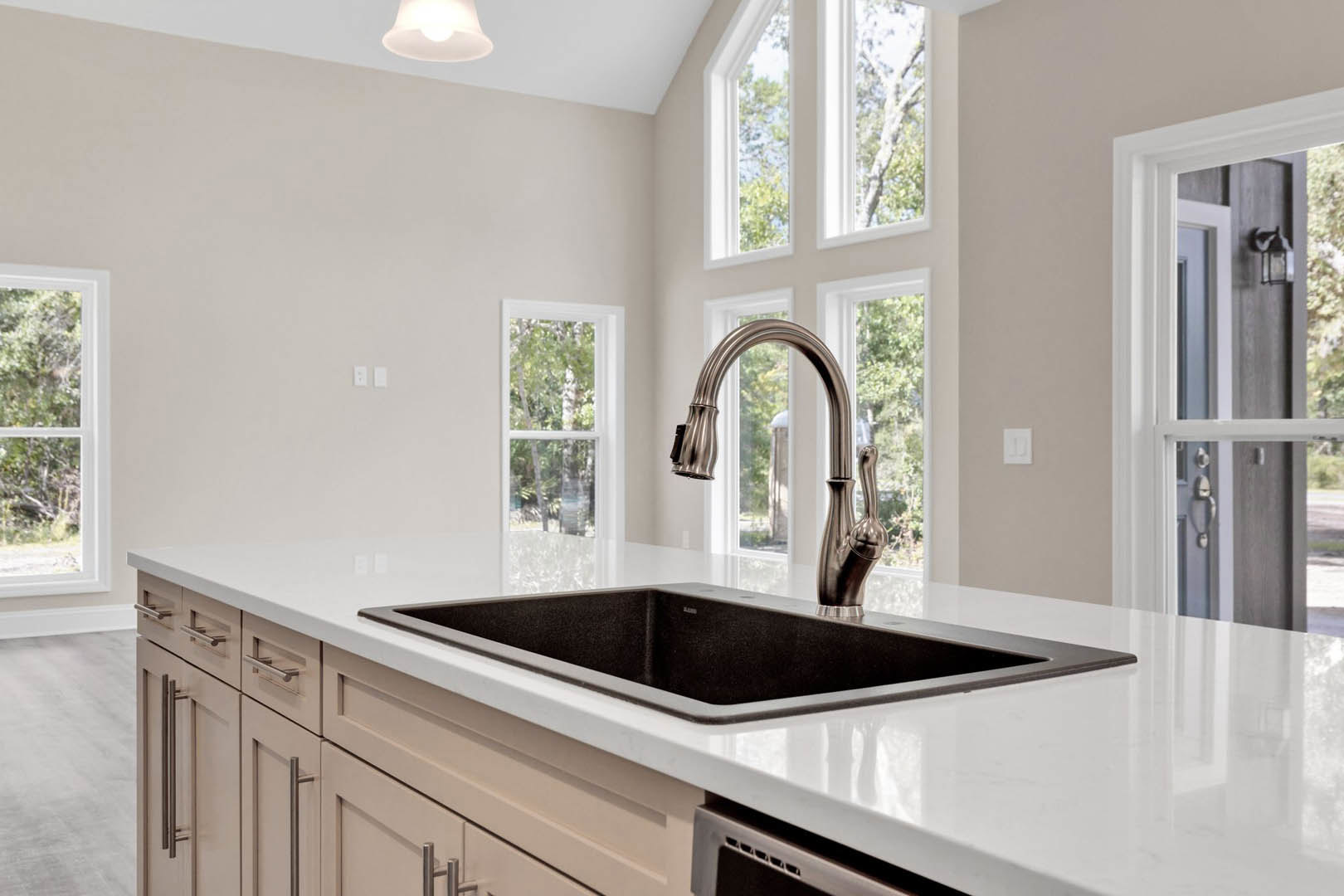 Modern kitchen with white countertops, stainless steel sink and curved faucet, black rectangular sponge in sink, upper cabinetry, large windows overlooking green trees, recessed