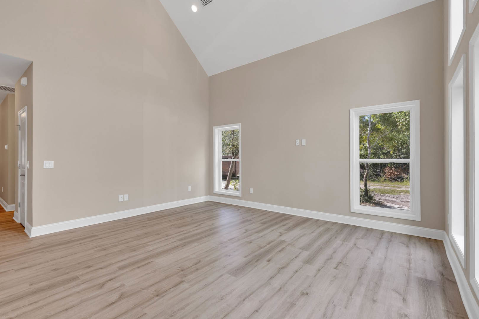 Sunlit room with wide plank hardwood flooring, large windows framing leafy trees, white plaster walls, and a simple ceiling light fixture