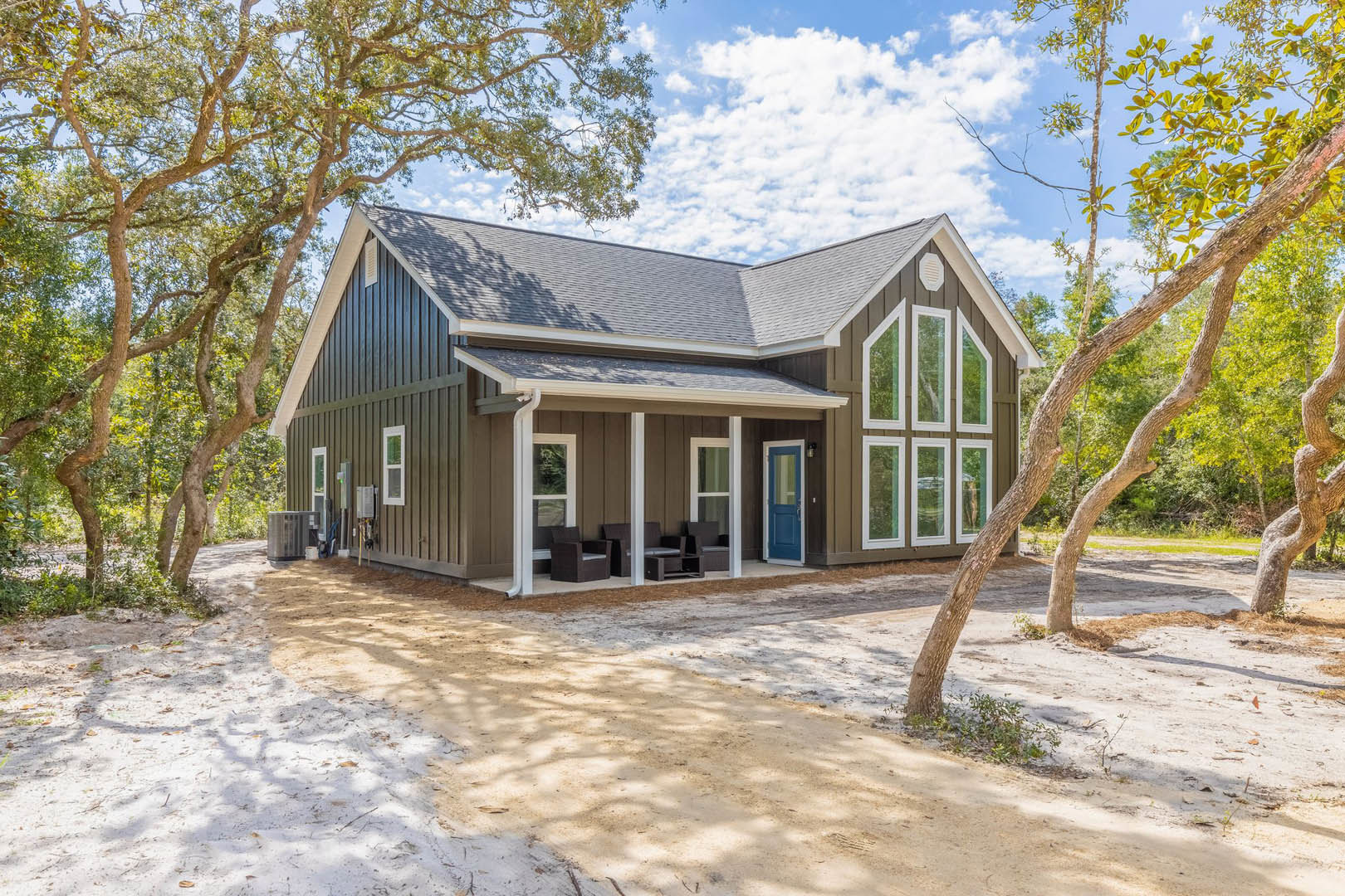 Two-story home with white siding, blue front door, white-framed windows, covered porch with chairs, surrounded by mature trees and dirt driveway