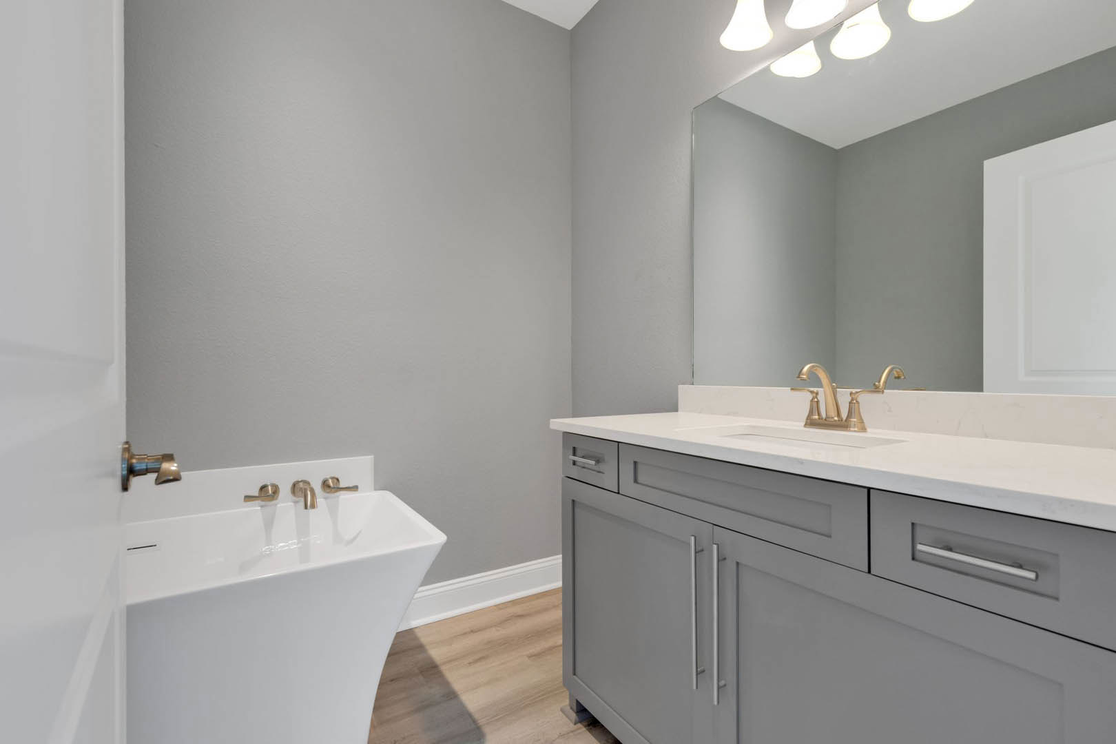 White bathroom vanity with gold faucet, rectangular mirror above sink, white cabinetry, wood flooring, and white door in background