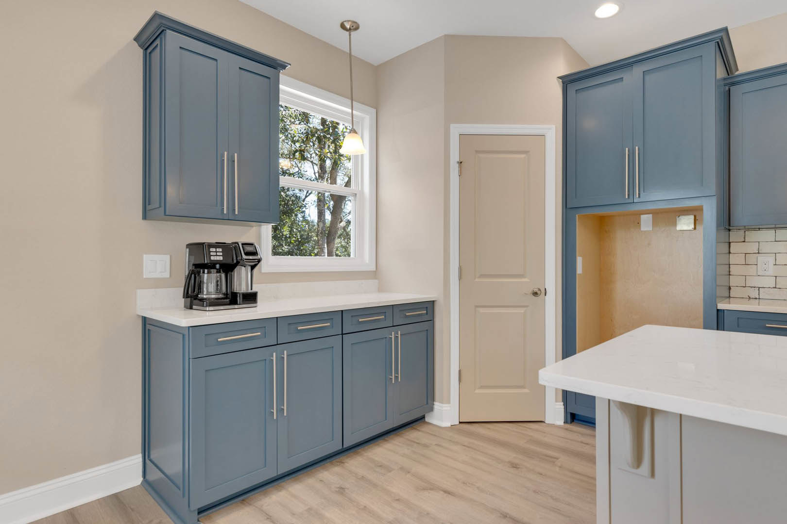 Blue shaker cabinets with brushed nickel hardware, white quartz countertop, stainless steel sink, coffee maker on counter, light streaming through window, white square wall outlet