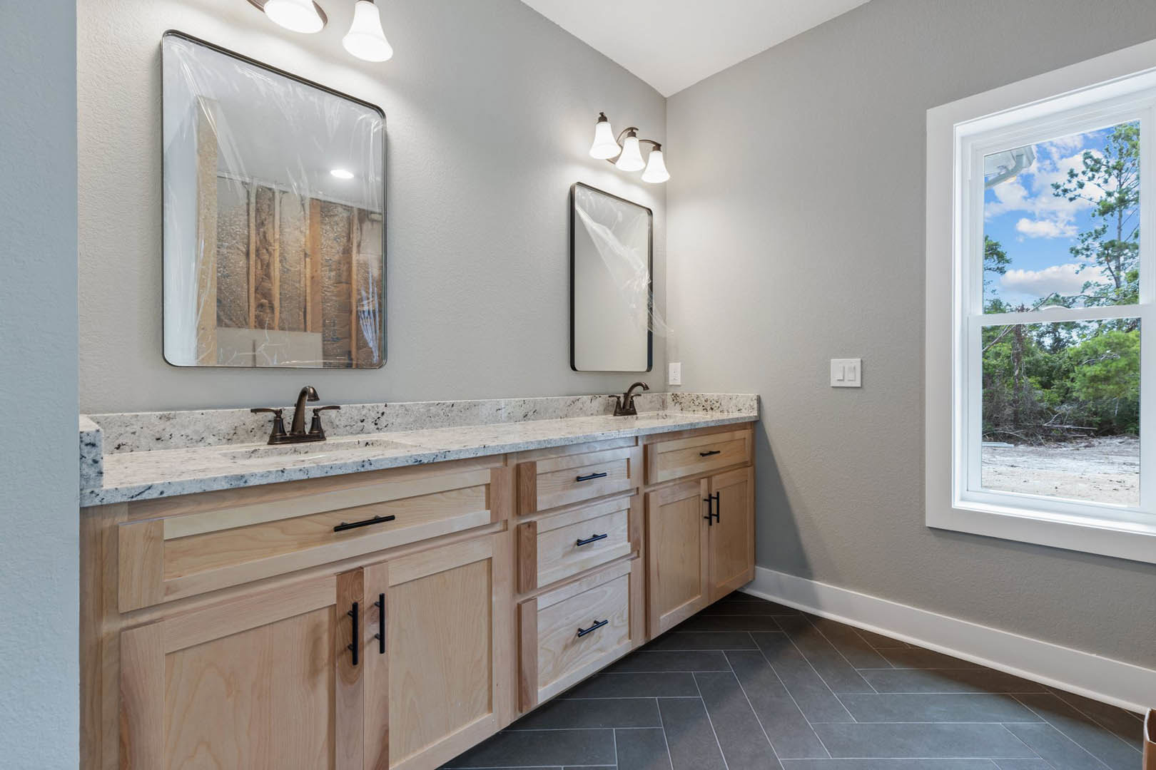 Bathroom with double sink vanity, large mirror, white countertop, cabinetry with drawers, tile flooring, plastic covering on wall, window showing trees, white light switch, plastic