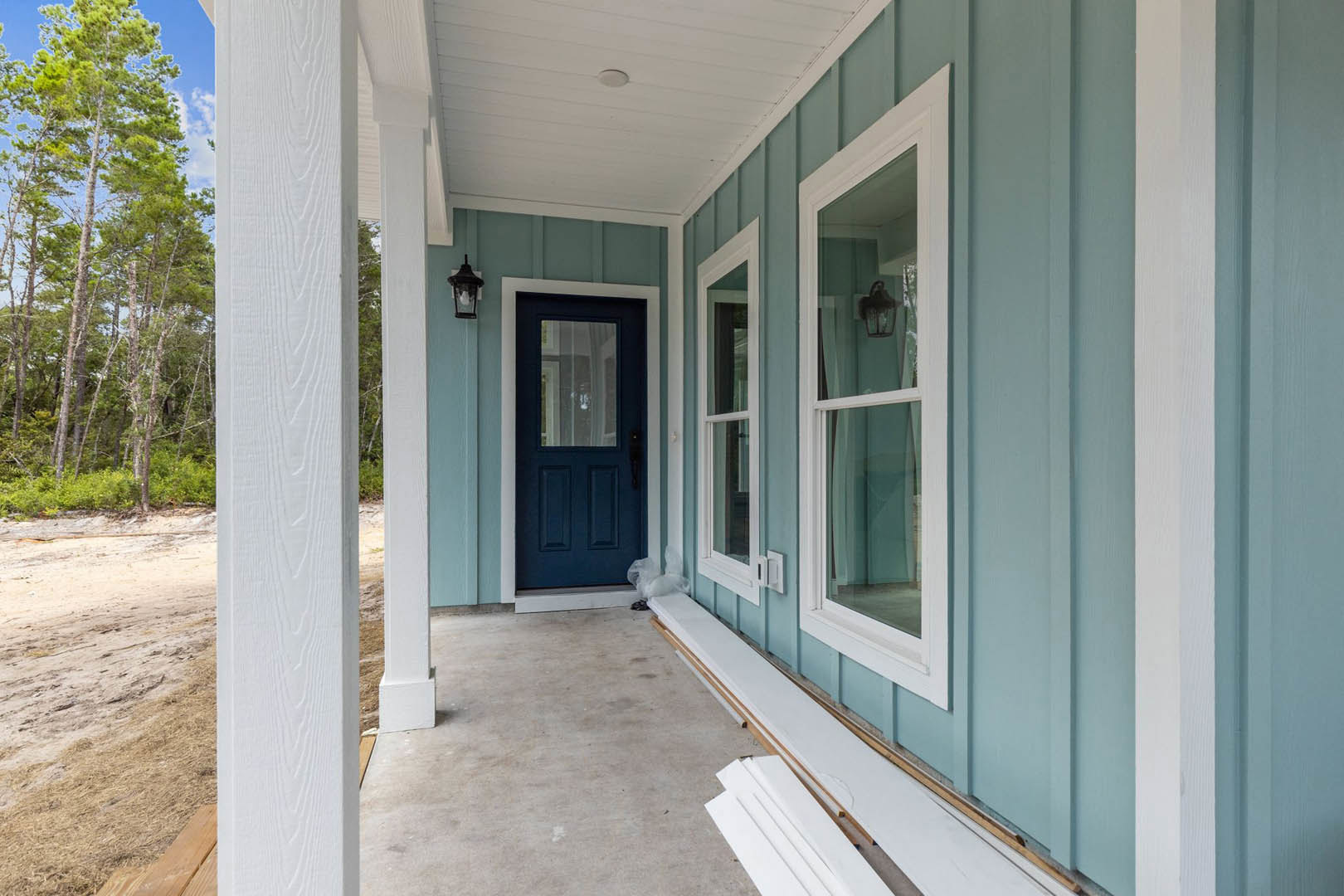 Blue siding house with white trim, blue front door with glass window, illuminated window, covered porch, mature trees in background, light sky above, composite porch flooring