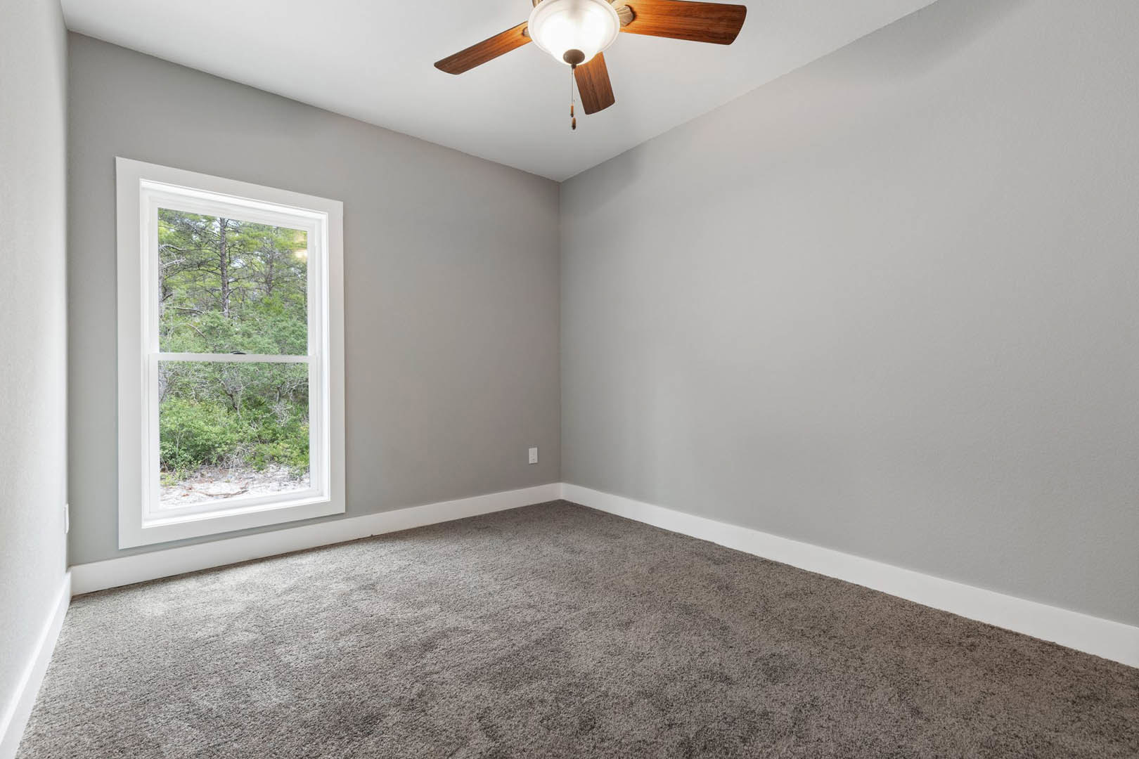 Neutral-toned room with carpet flooring, white plaster walls, large window overlooking trees, and ceiling fan with integrated light fixture