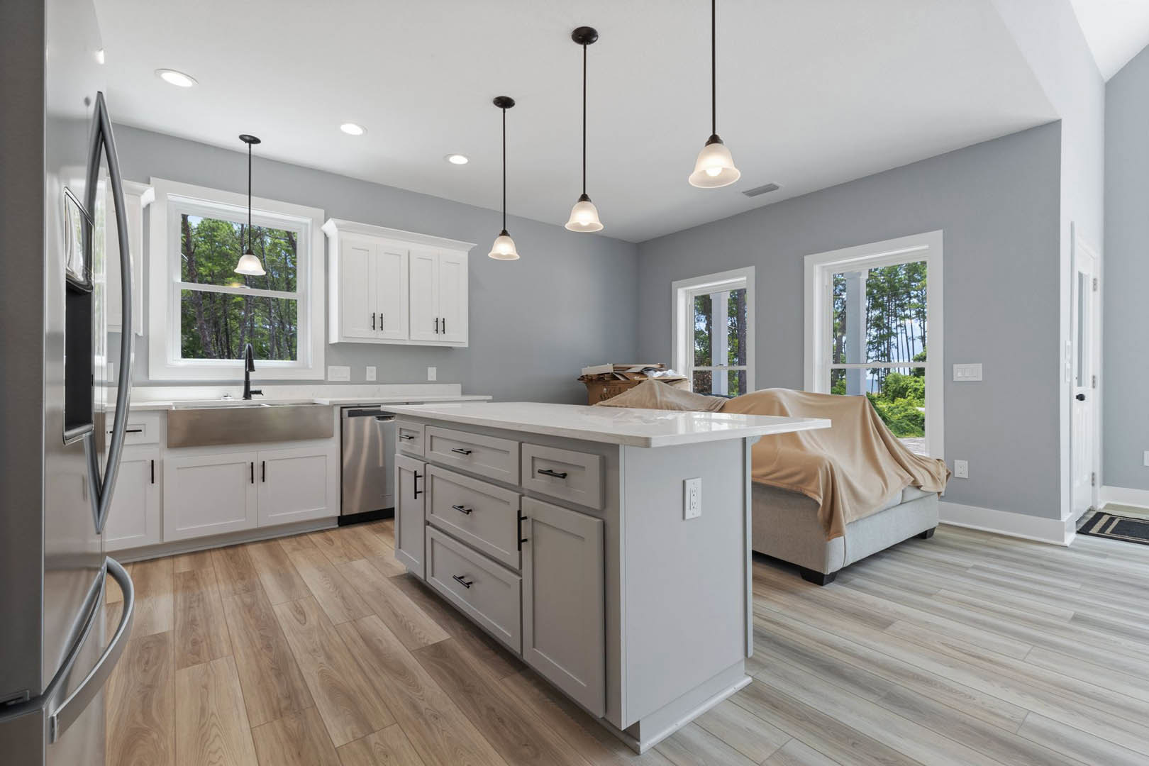 White kitchen with wood flooring, white cabinets and island featuring black handles, stainless steel refrigerator, sink beneath window, pendant lights hanging from ceiling