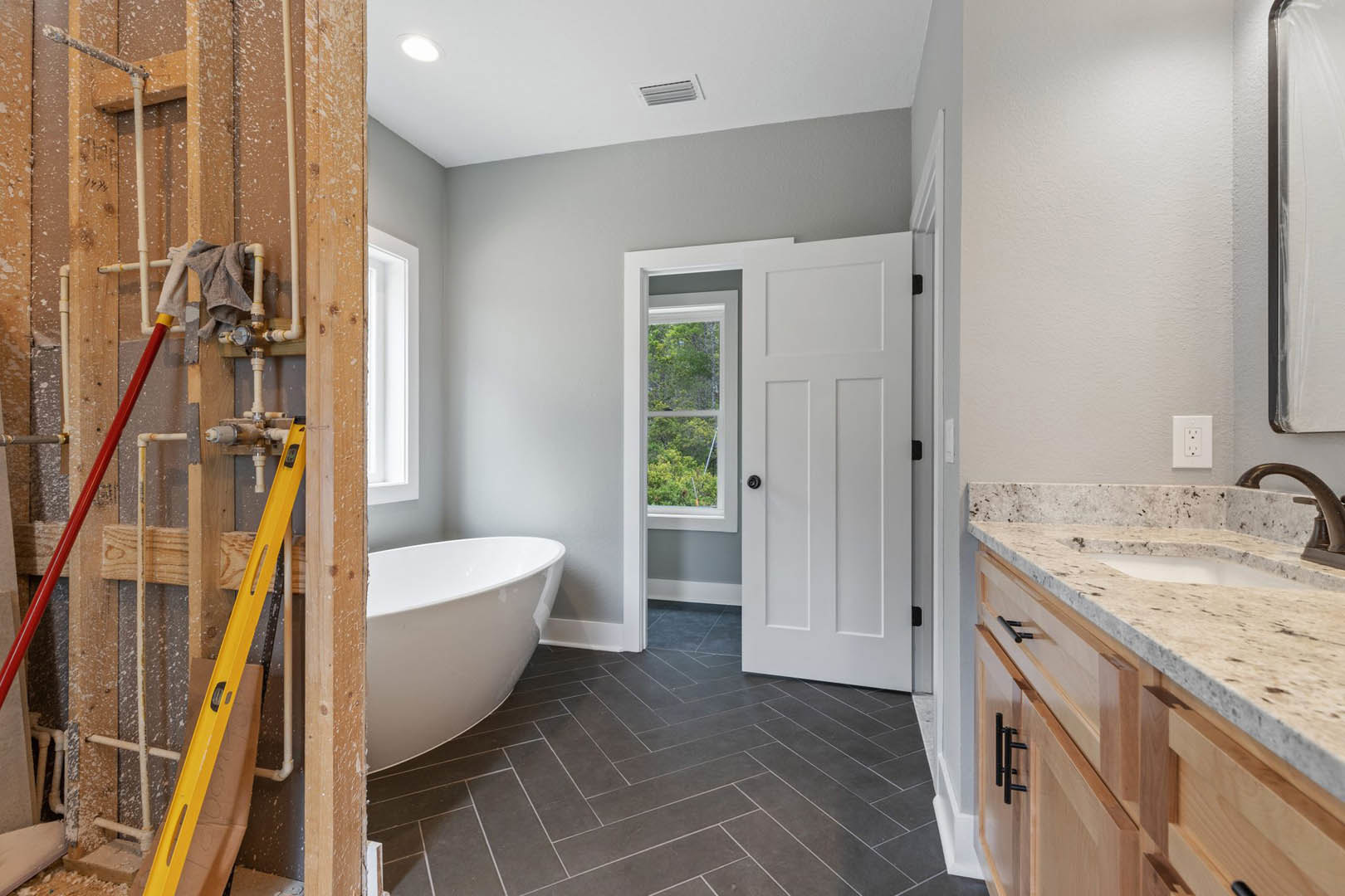 Bathroom with freestanding white bathtub, rectangular sink on wood vanity, grey tile floor with white grout lines, large window showing trees, white door with glass panel, white