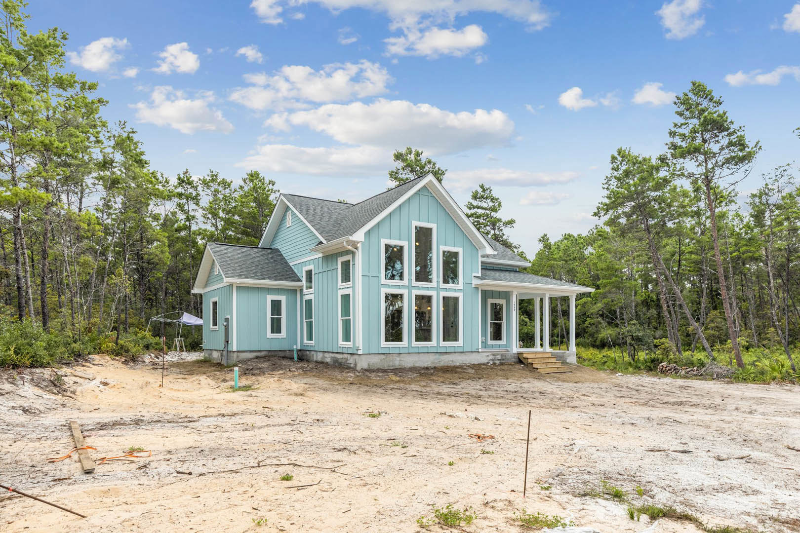 Blue house with white trim surrounded by tall trees, unfinished dirt yard, covered front porch, staircase leading to entrance, partly cloudy sky overhead