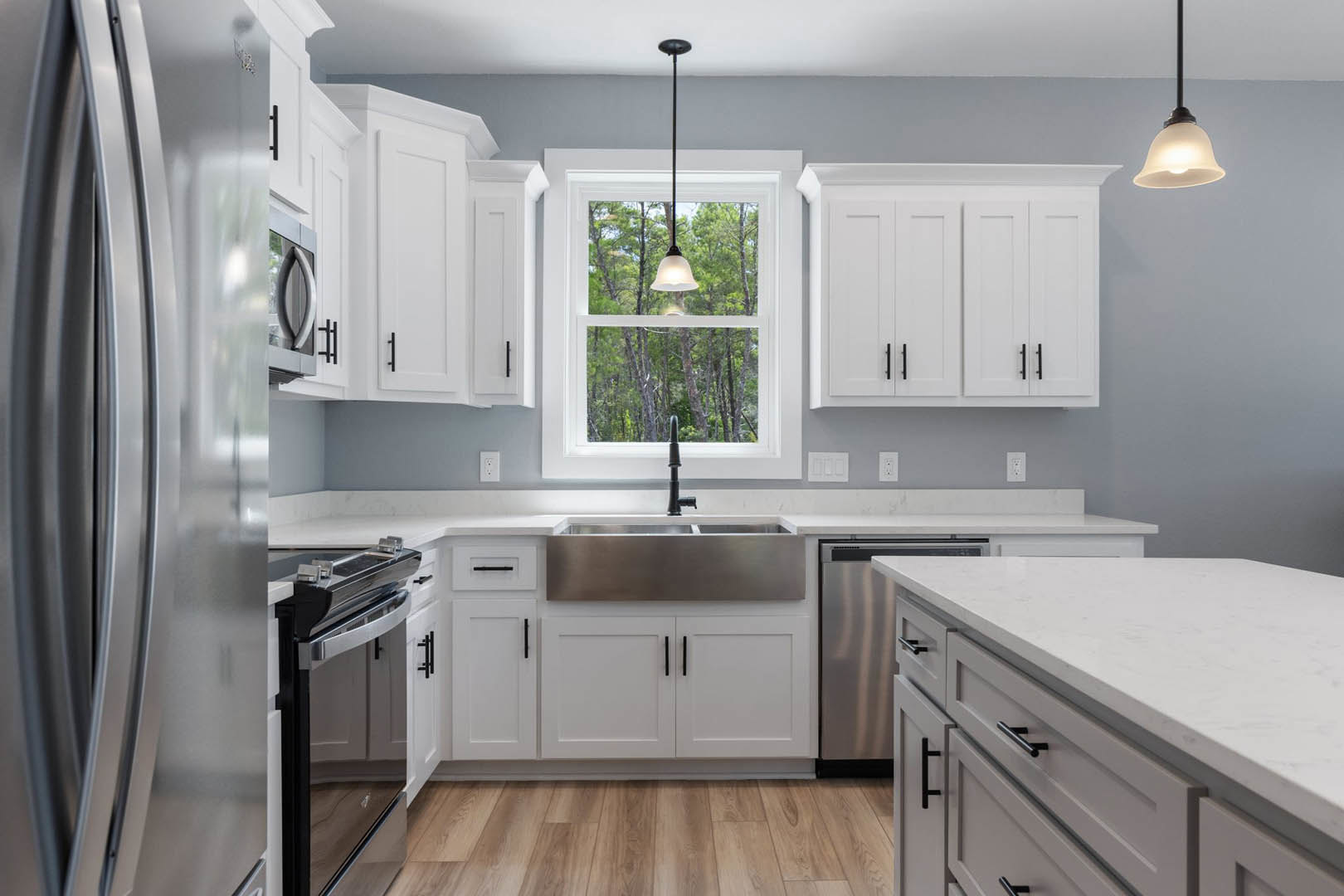 White kitchen with shaker cabinets and black handles, wood flooring, central island, pendant light fixture, large window above sink, stainless steel appliances.