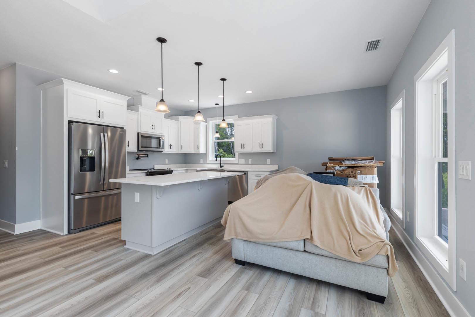 Open kitchen with white island, wood flooring, stainless refrigerator, built-in microwave with black handle, ceiling vent, and a couch draped in a blanket.