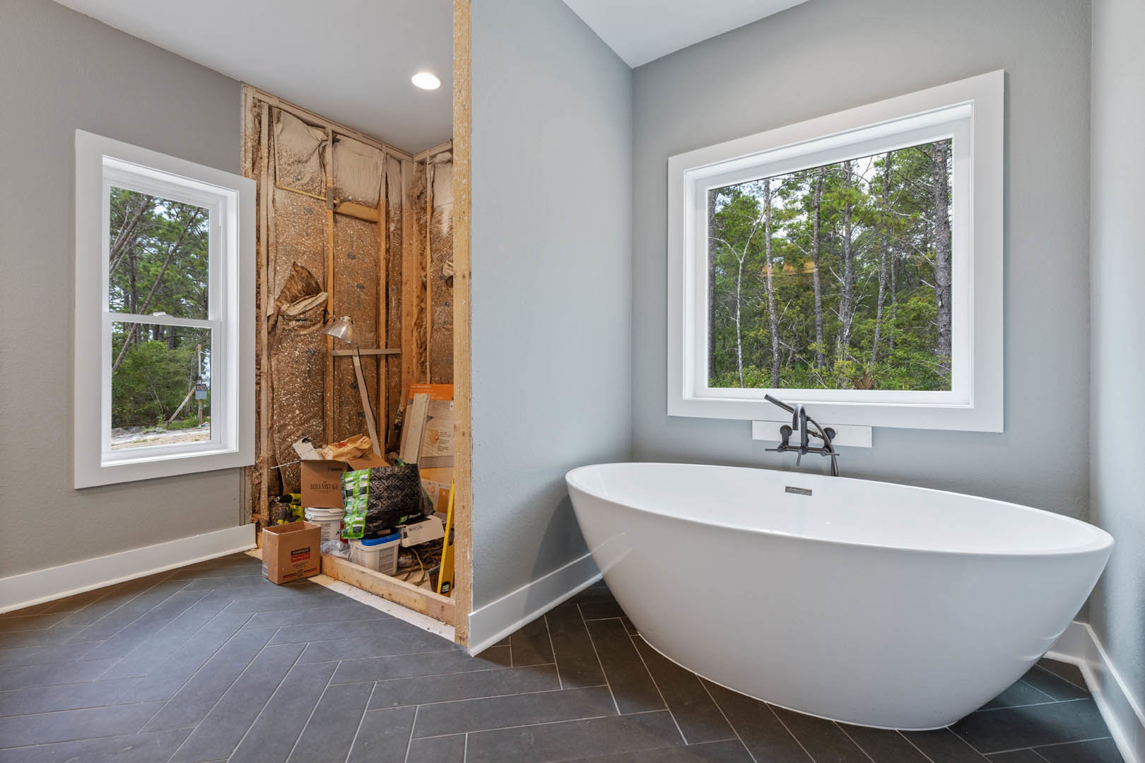 White freestanding bathtub with chrome faucet and hose, large window overlooking trees, cardboard box and white box with black text on countertop, light tile walls and floor