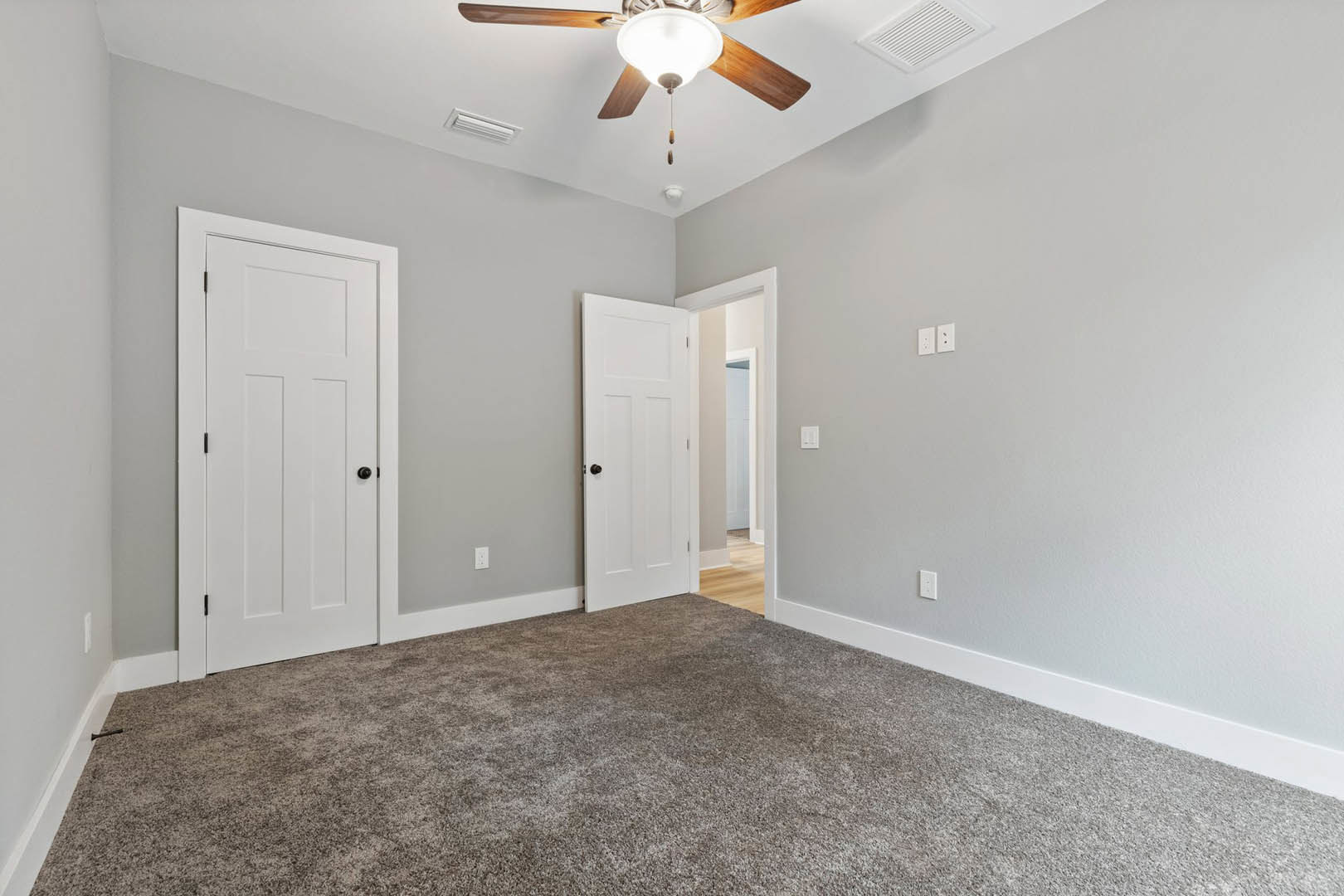 Carpeted room with white paneled doors featuring black knobs, ceiling fan with light, white wall outlet, and white vent on smooth plaster walls and ceiling.