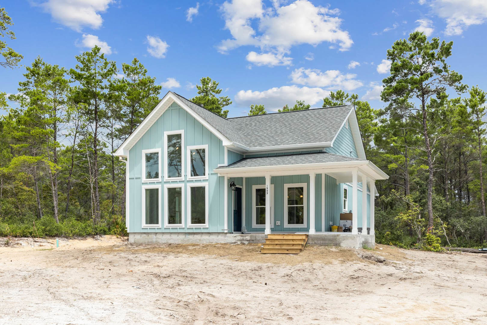 Blue house with white trim and windows, wooden fence in front, mature trees and partly cloudy sky in background