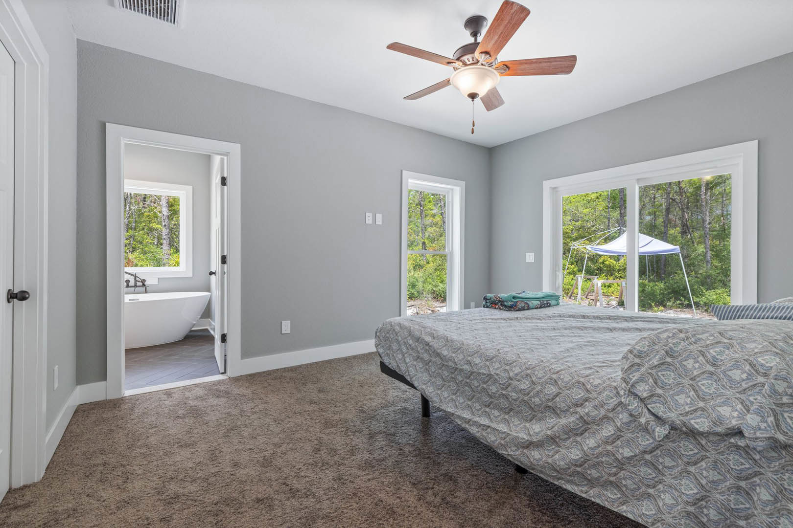Bedroom with wood floor, ceiling fan with light, bed with white blanket, large window overlooking trees, freestanding white bathtub in adjacent bathroom.
