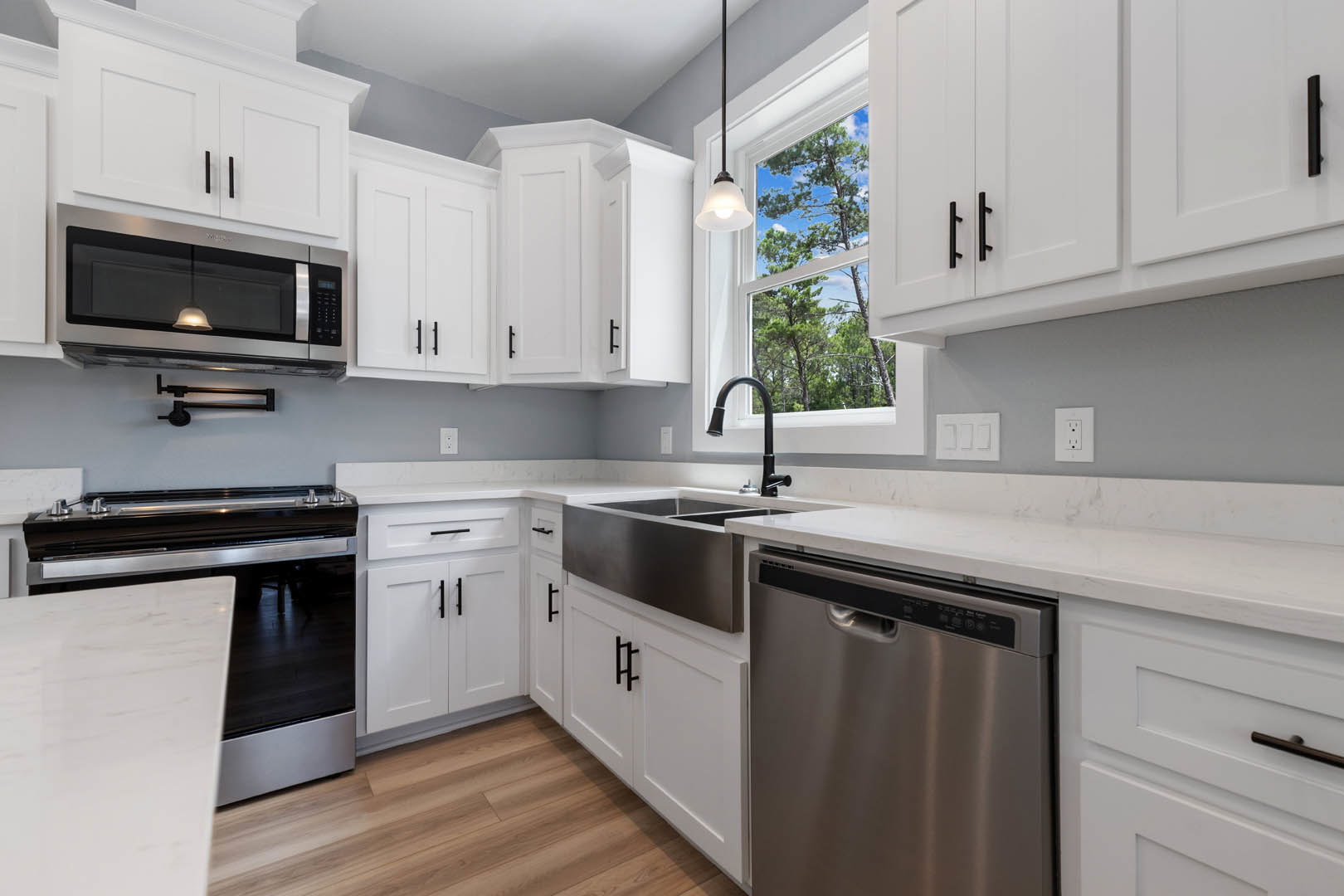 White kitchen cabinets, stainless steel appliances, light fixture hanging from ceiling, dishwasher with handle, white table beside black and silver oven, white electrical outlet