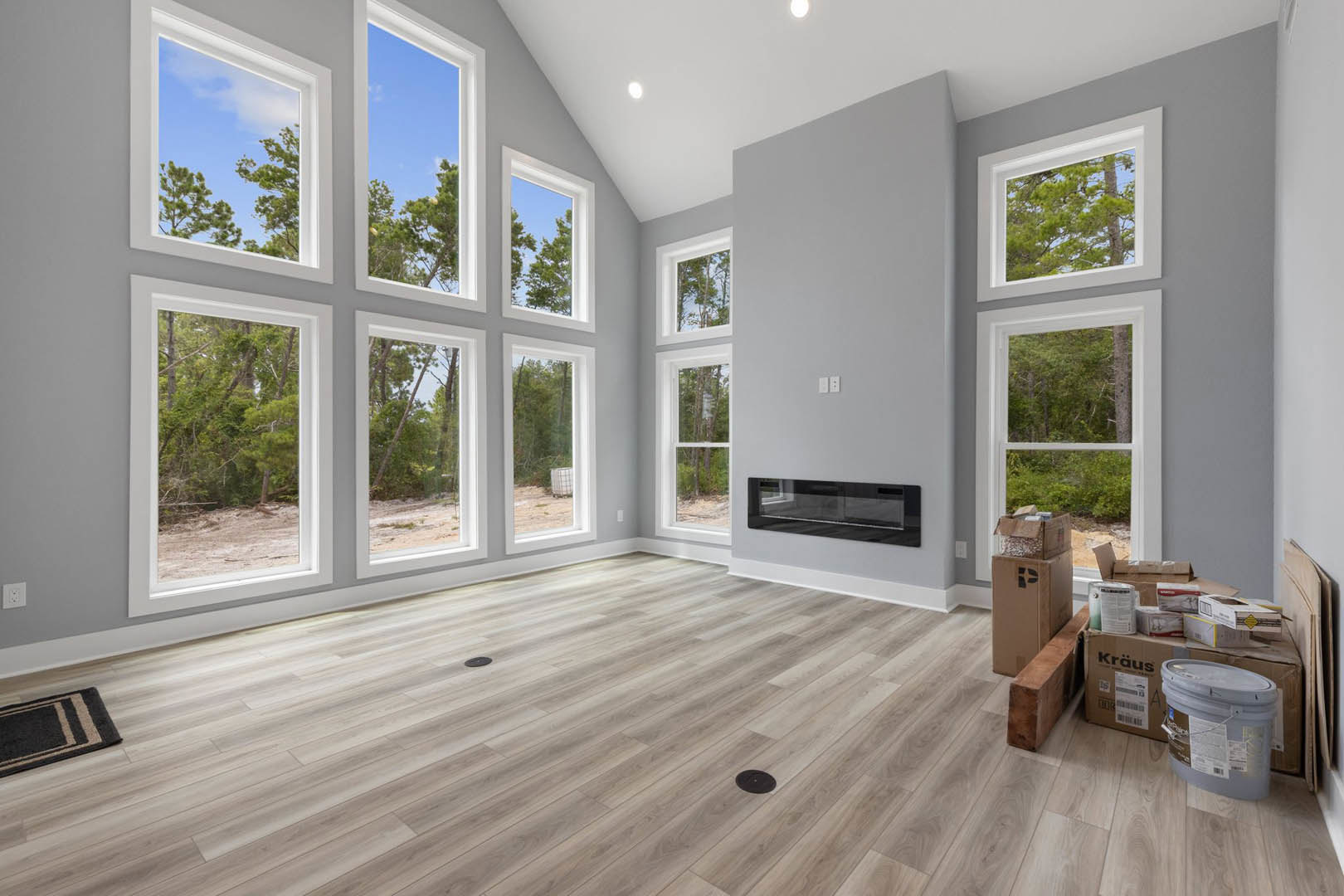 Living room with wood flooring, black and white rug, multiple large windows offering forest views, and a stone fireplace