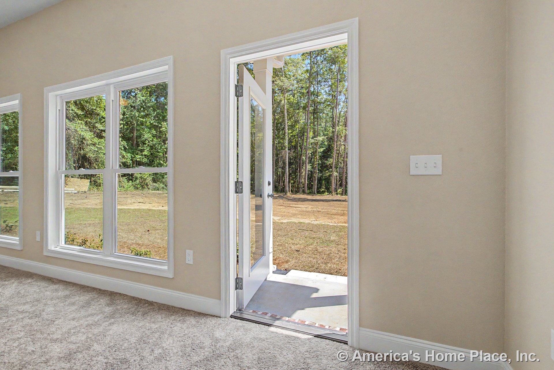 Carpeted room with beige walls, large double windows with white trim, glass door opening to covered patio, white baseboards, and wall-mounted light switches.