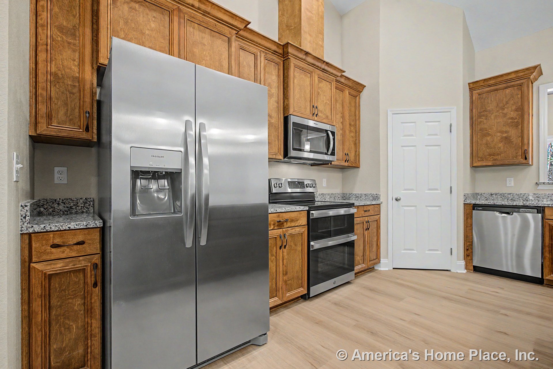 Wood upper and lower kitchen cabinets with granite countertops, stainless steel refrigerator, oven, microwave, and dishwasher, light wood flooring, and a white paneled door.