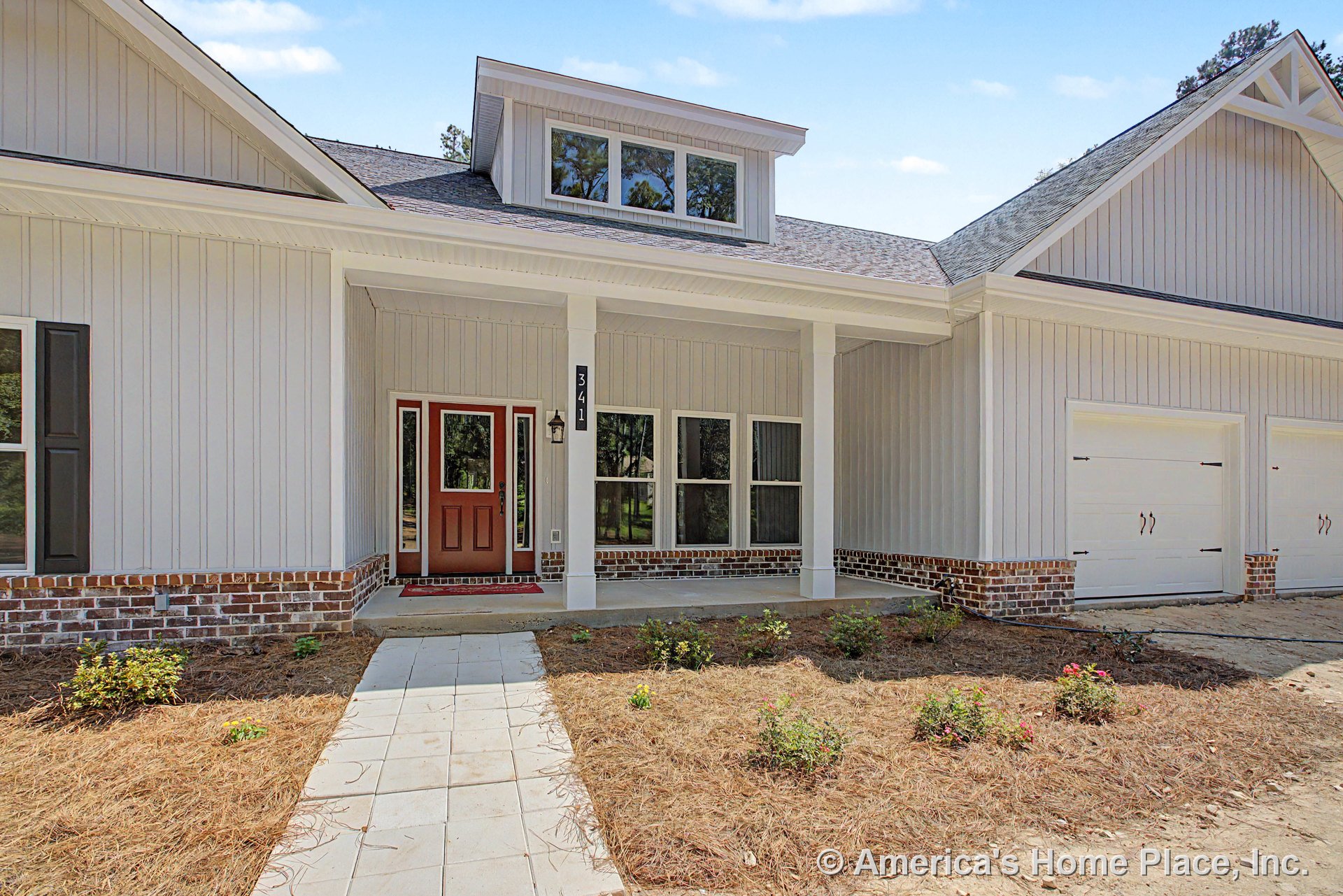 Vertical board siding with brick foundation trim, covered front porch supported by columns, double garage doors, multi-pane windows, red entry door with glass panels, roof dormer