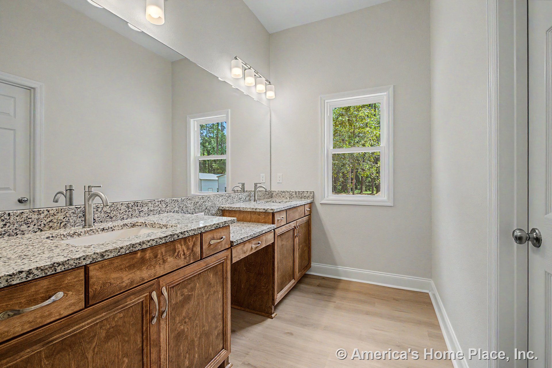 Dual undermount sinks set in granite countertops with wood cabinetry, large wall mirror, modern vanity lighting, vinyl plank flooring, window with trim, neutral walls, and door.