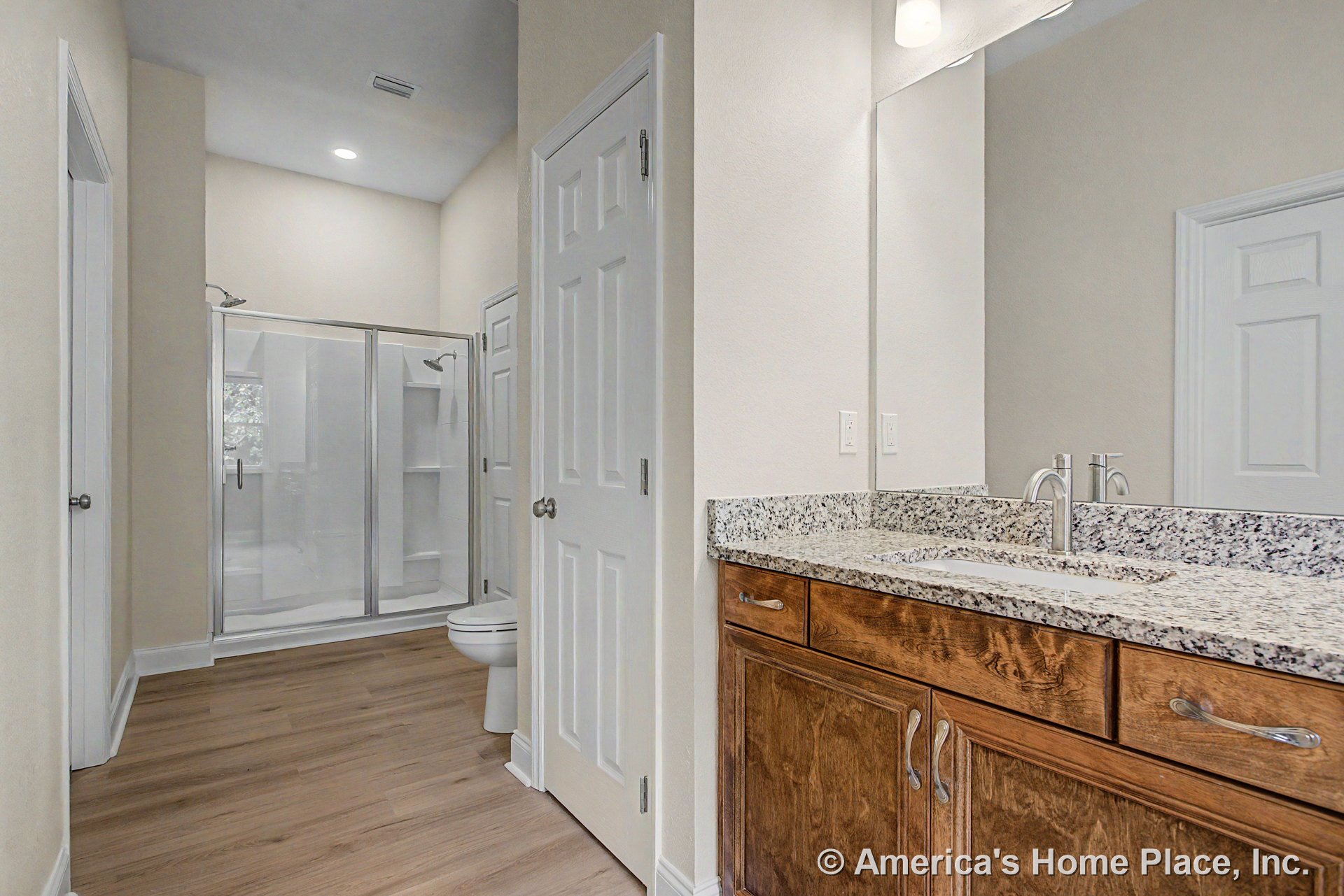 Double shower enclosure with clear glass doors, granite vanity countertop, wood cabinetry, expansive wall mirror, recessed ceiling lighting, and light wood flooring.