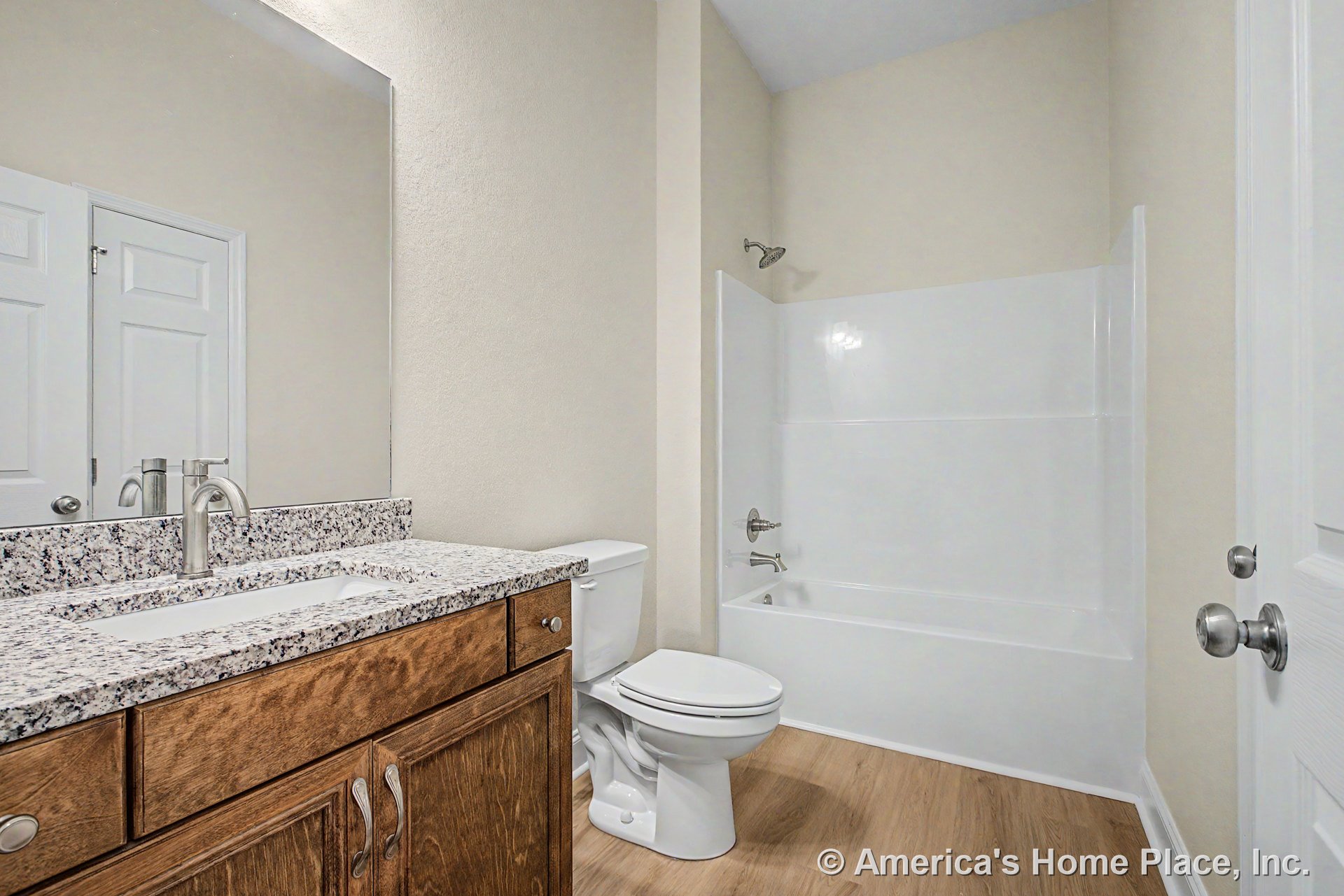 Granite countertop atop wood vanity with single lever faucet, fiberglass tub-shower combo, light wood-look flooring, white paneled doors, and neutral walls.