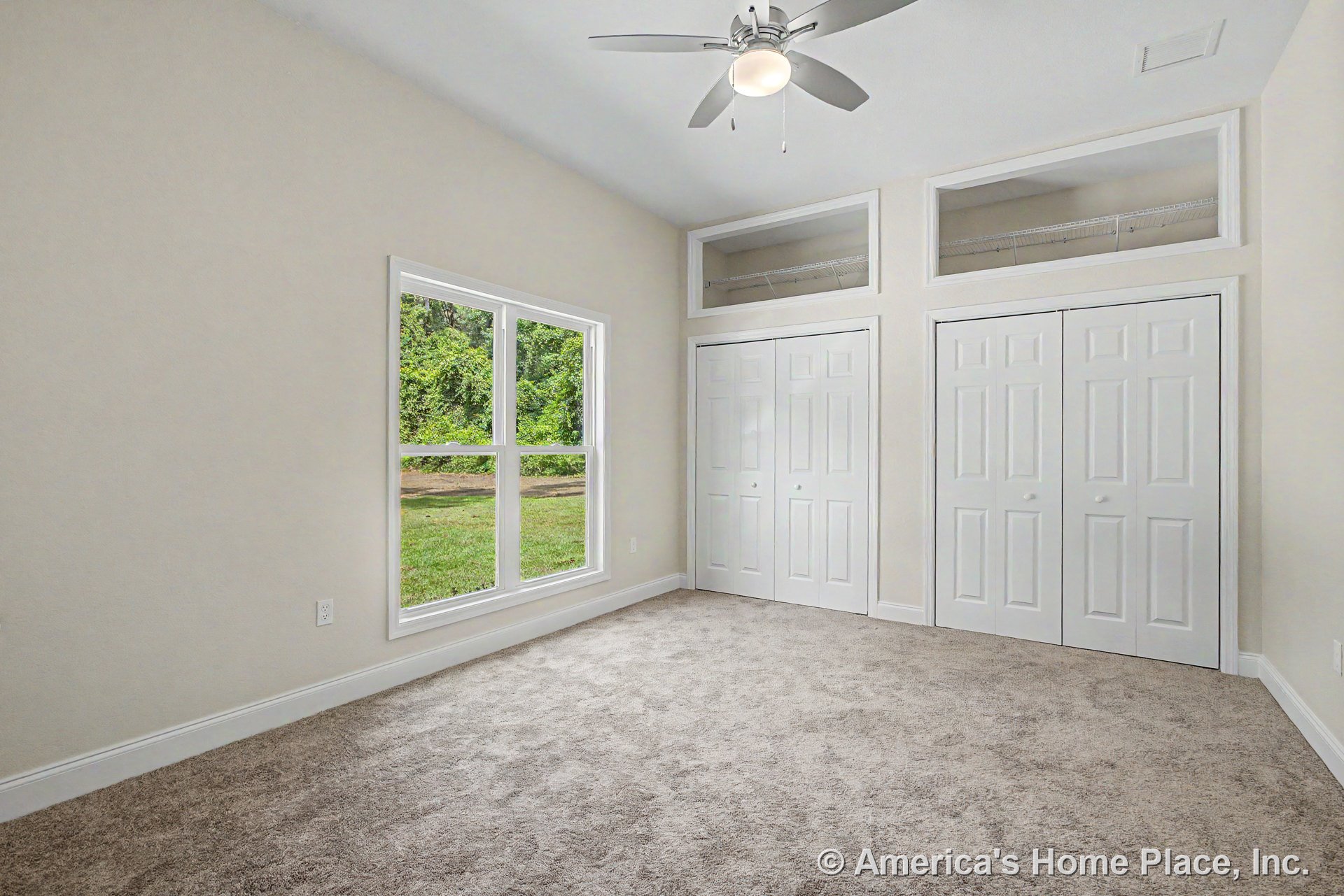 Bedroom with double closet doors topped by transom windows, large window offering outdoor view, neutral carpet flooring, ceiling fan with built-in light, white trim and baseboards