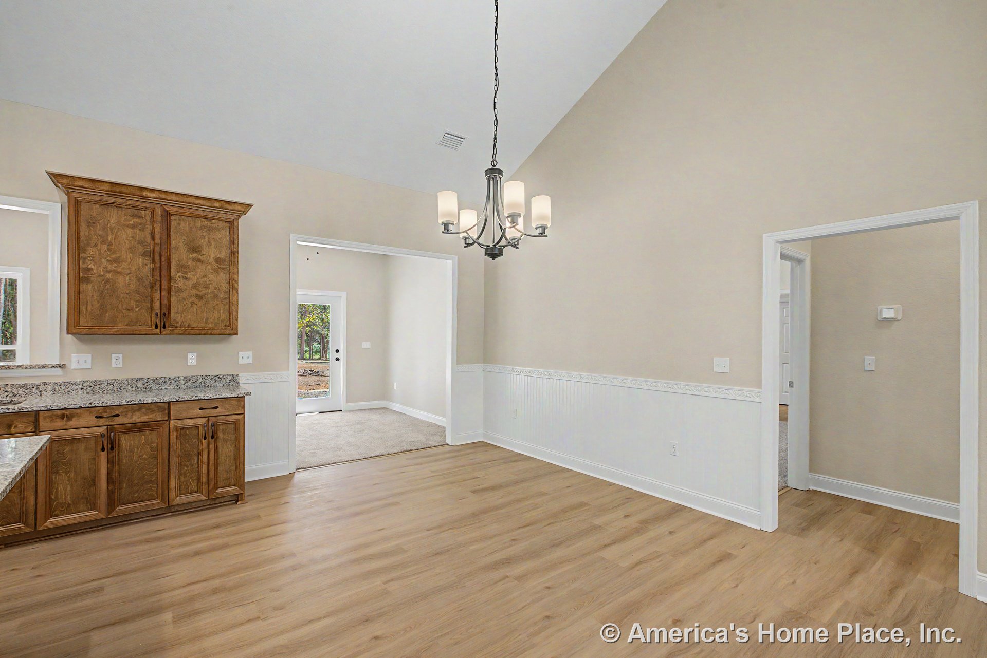 Vaulted ceiling dining area with wood-look flooring, five-light chandelier, wainscoting and trim, granite countertops, wood cabinetry, neutral wall color, and open layout.