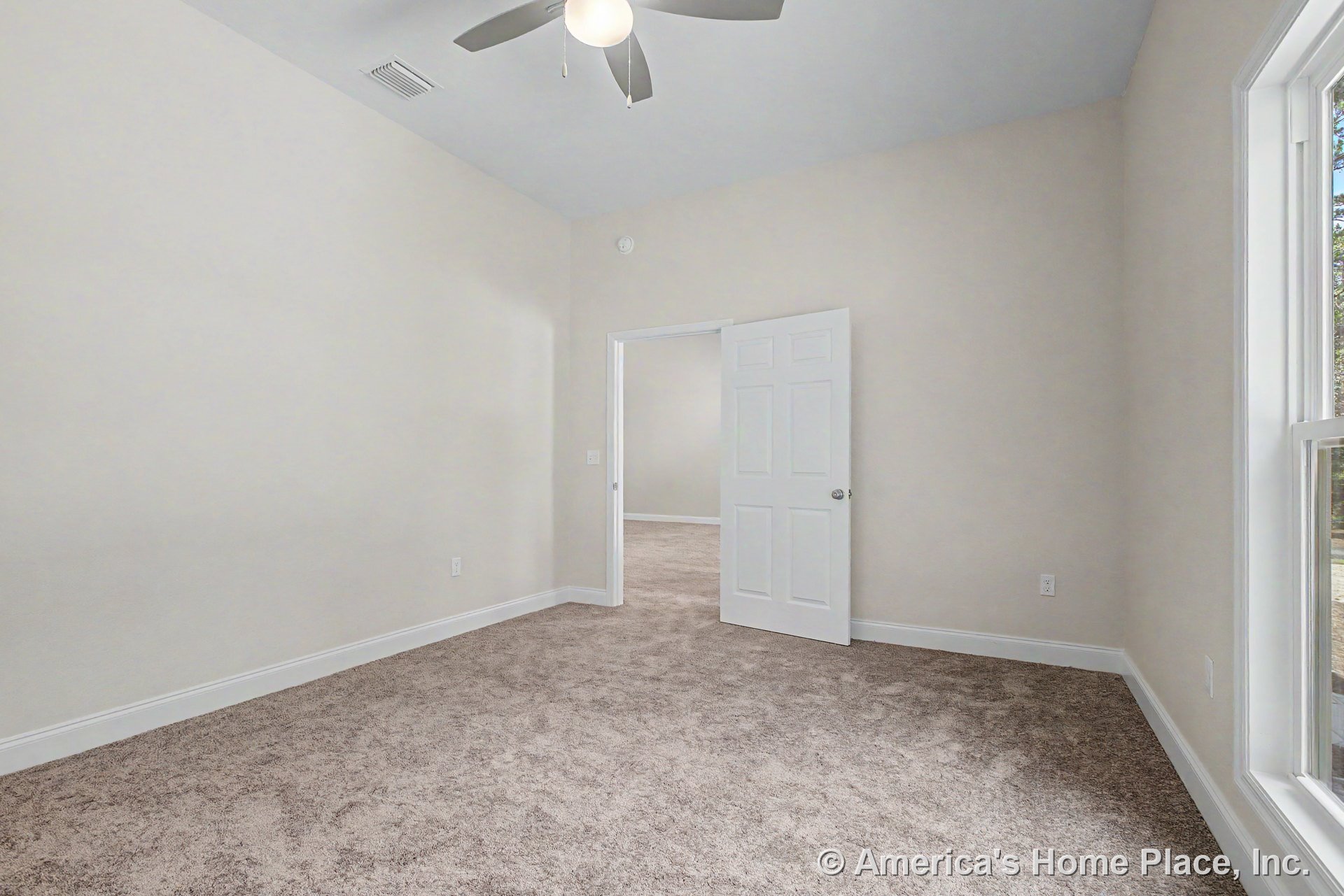 Bedroom with beige carpet flooring, white trim and baseboards, ceiling fan with light fixture, large window allowing natural light, white paneled door, and neutral wall color.