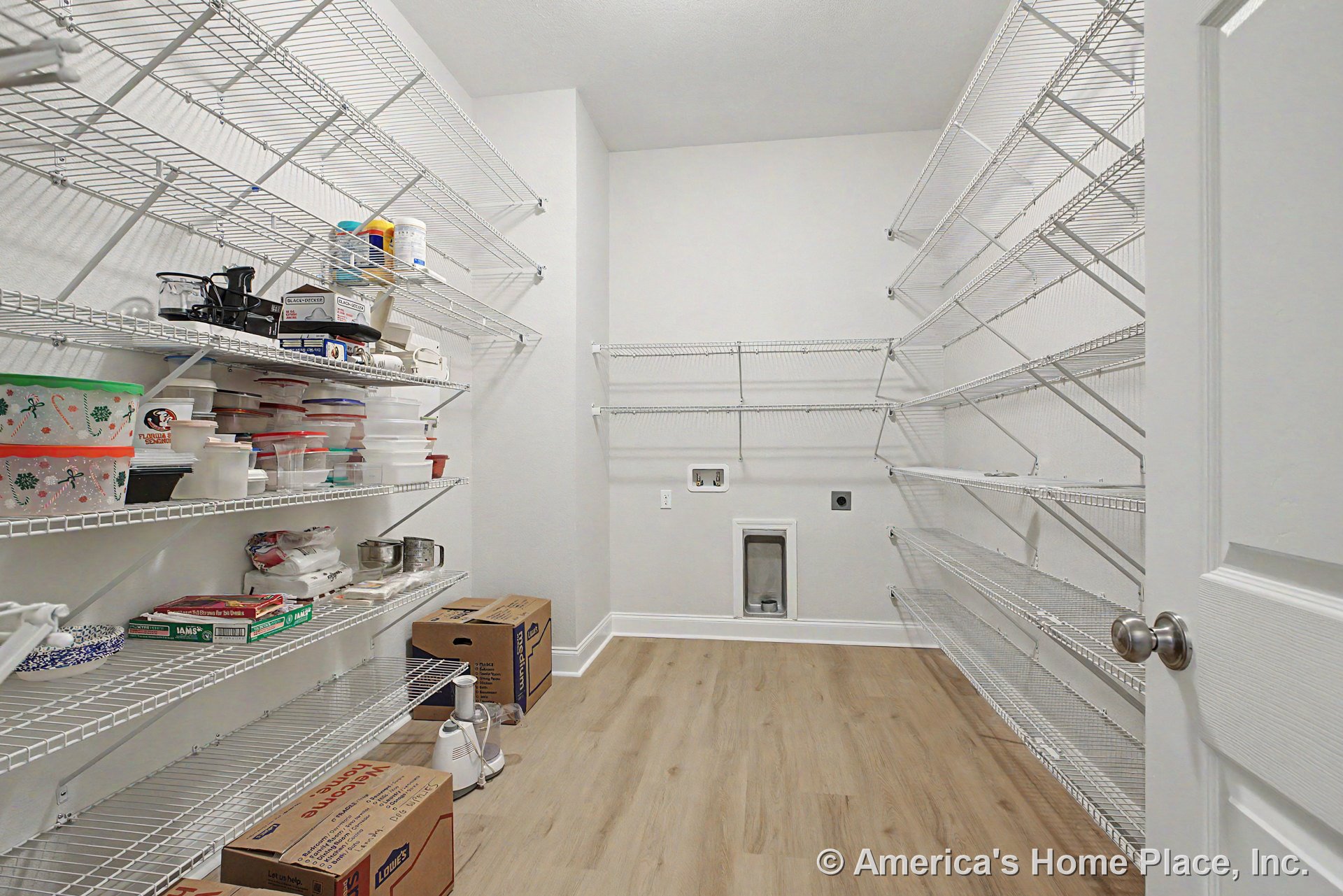 Walk-in pantry with extensive built-in wire shelving, light wood flooring, white painted walls, utility hookups, white door with knob, and baseboard trim.
