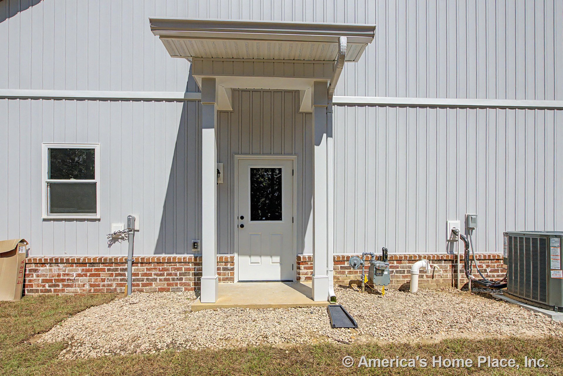Covered entry porch with white paneled door set in vertical siding, brick foundation trim, single window, visible HVAC unit and utility meters, and concrete step.