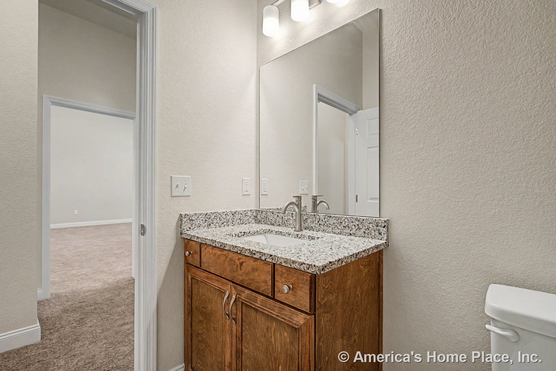 Granite countertop vanity with wood cabinetry, large wall mirror, built-in overhead lighting, bathroom sink and faucet, neutral wall paint, white trim, doorway, and toilet.